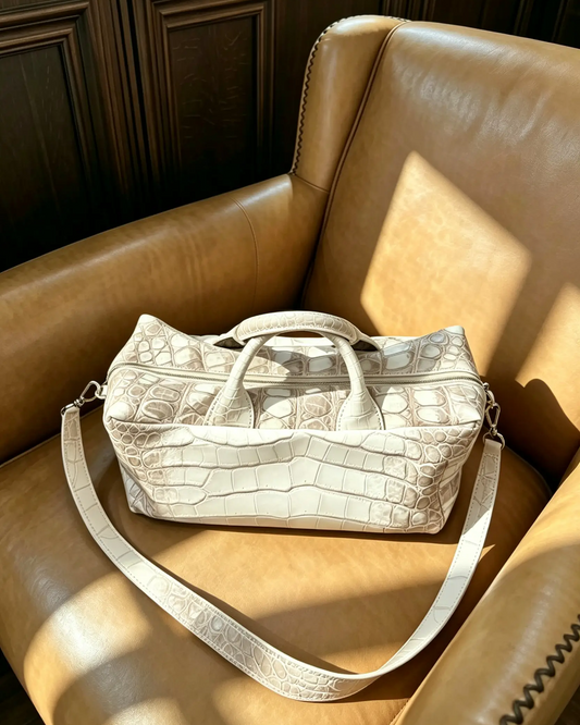 A light beige crocodile-embossed duffel bag with a matching shoulder strap, resting on a brown leather armchair in warm sunlight.