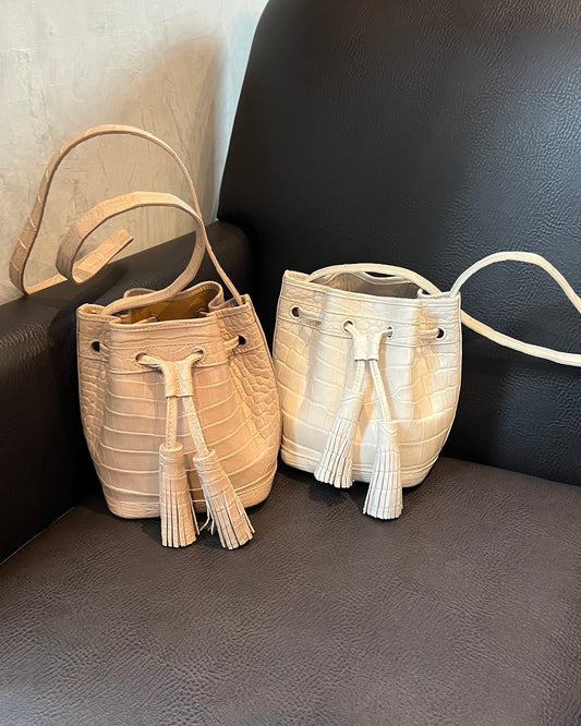 Two small bucket-style handbags with tassel drawstrings and textured leather-like surfaces, placed on a dark upholstered chair. One bag is a light cream/off-white color and the other is a warm beige/tan color.