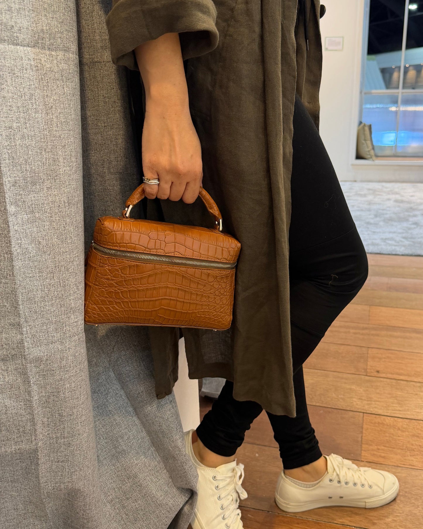Person sitting on a couch holding a brown leather bag, wearing white sneakers and a long coat.
