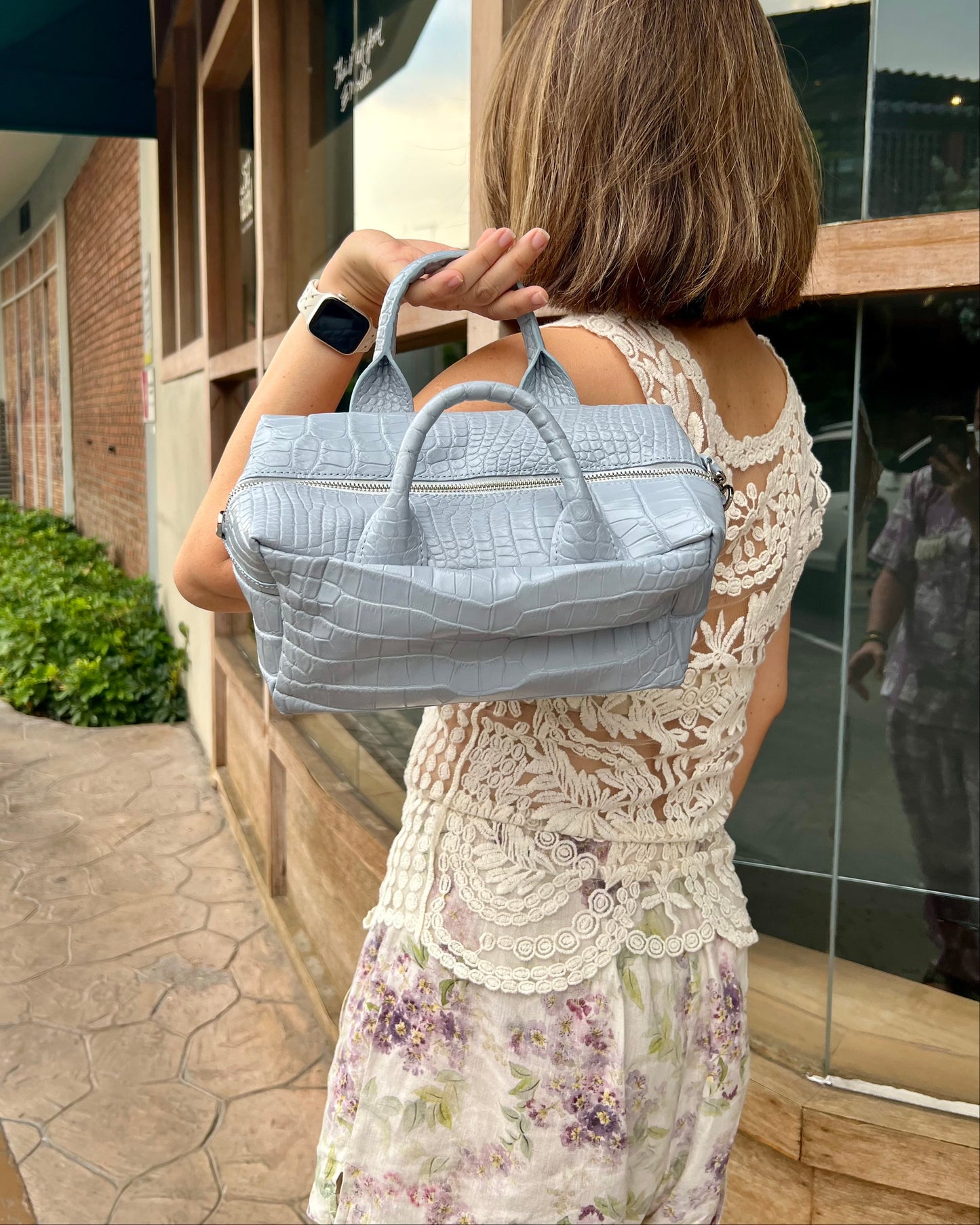 A woman viewed from behind carries a light blue crocodile-embossed handbag over her shoulder while wearing a cream lace dress on an outdoor walkway.