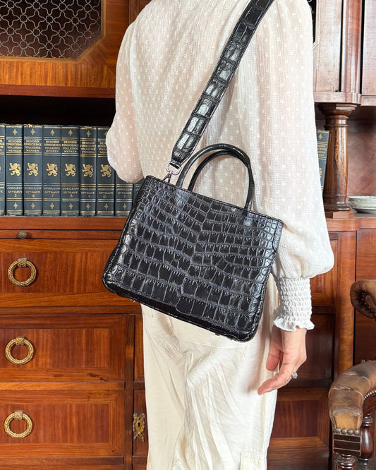 A woman viewed from behind wearing a textured cream dress and carrying a black crocodile-embossed leather shoulder bag with a top handle, standing in front of a large wooden bookcase.