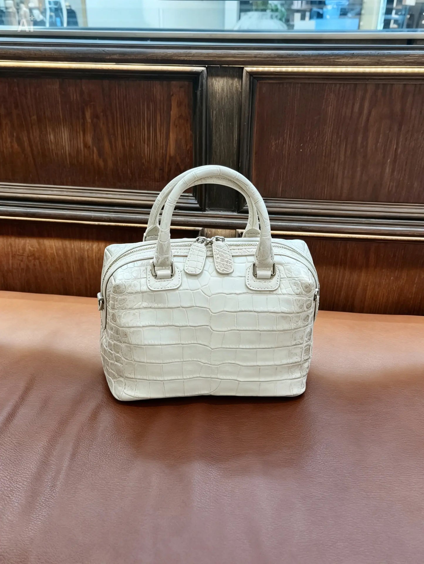 A structured white crocodile-embossed handbag with double top handles, resting on a brown leather surface in front of a dark wood-paneled background.