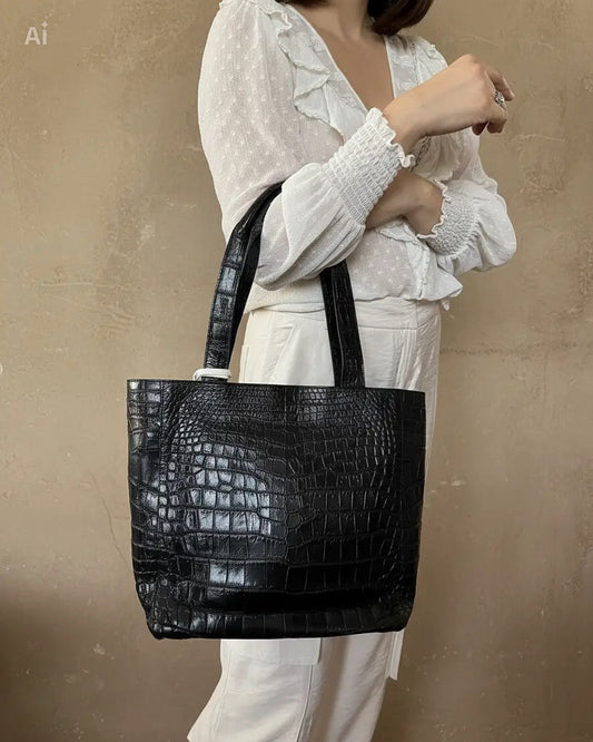 A person wearing a white blouse and white pants holds a large black crocodile-embossed tote bag against a textured beige wall.