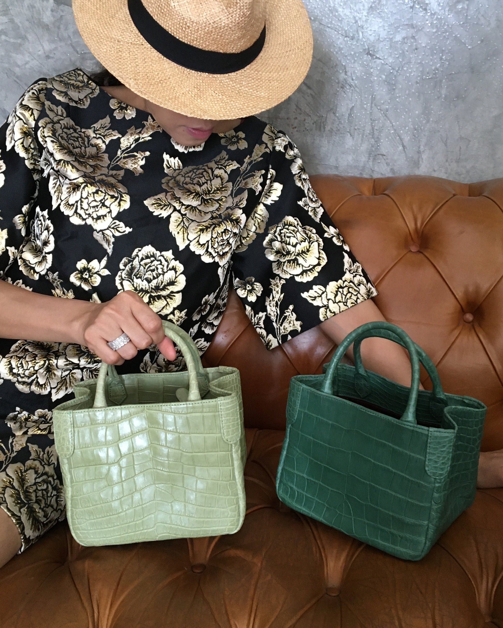 Person holding a green handbag on a brown leather couch with floral-patterned pants and a straw hat.