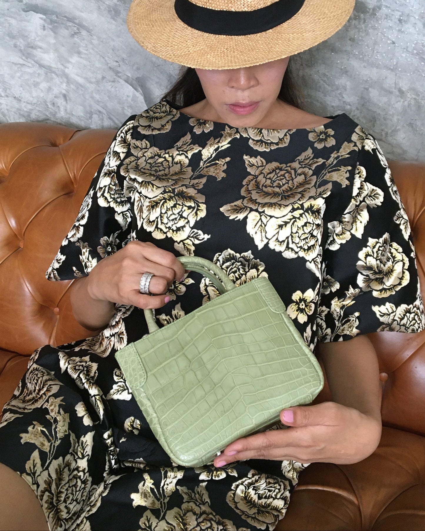Person sitting on a brown leather couch holding a green clutch and wearing a black floral dress.