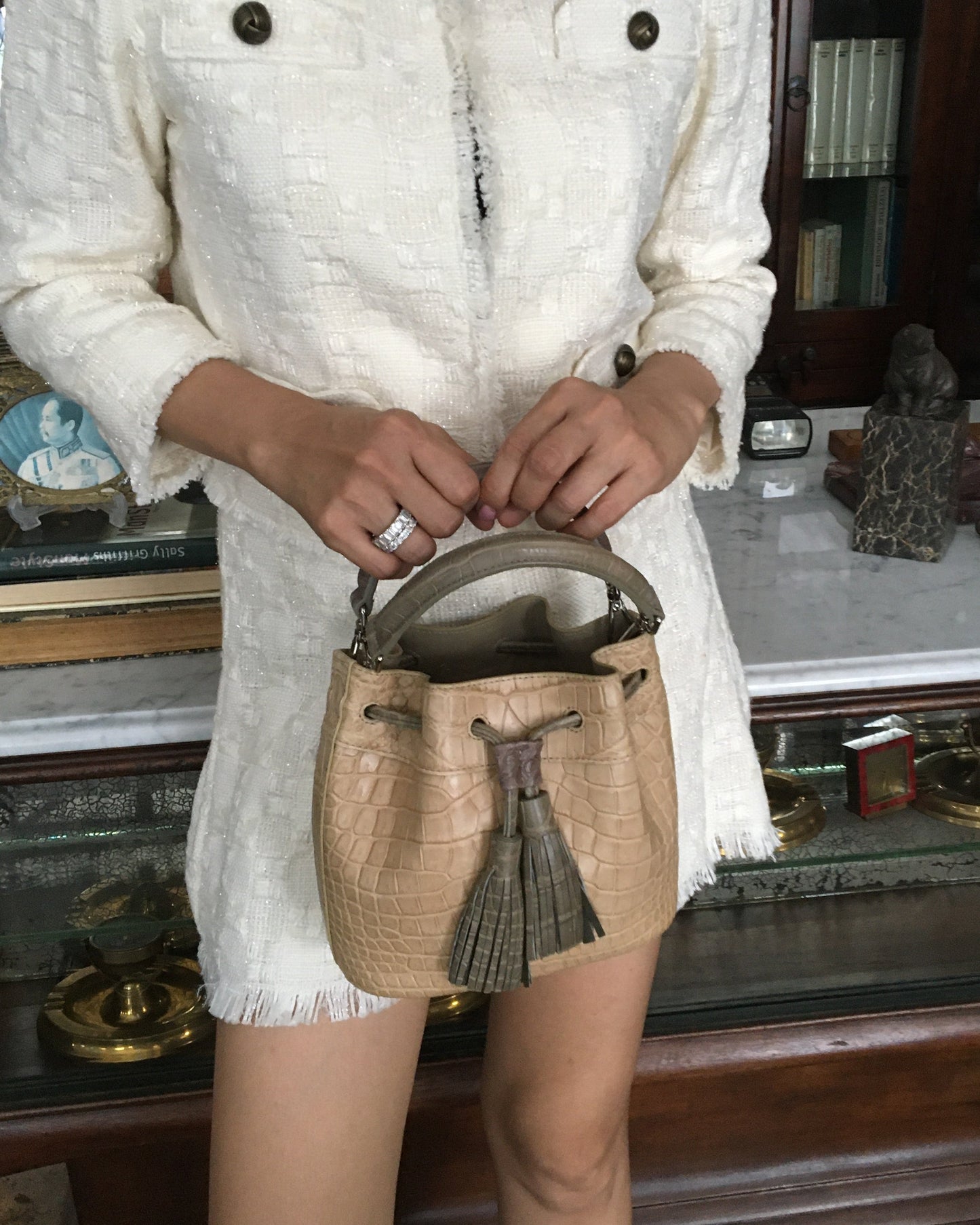 Person holding a beige handbag with a tassel in front of a white dress.