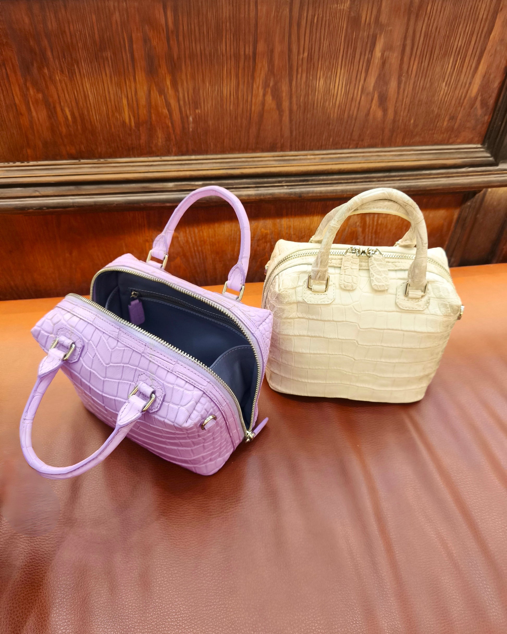 Two small structured handbags with crocodile-embossed texture, one lavender purple and one cream beige, displayed on a brown leather surface in front of a wooden panel background.