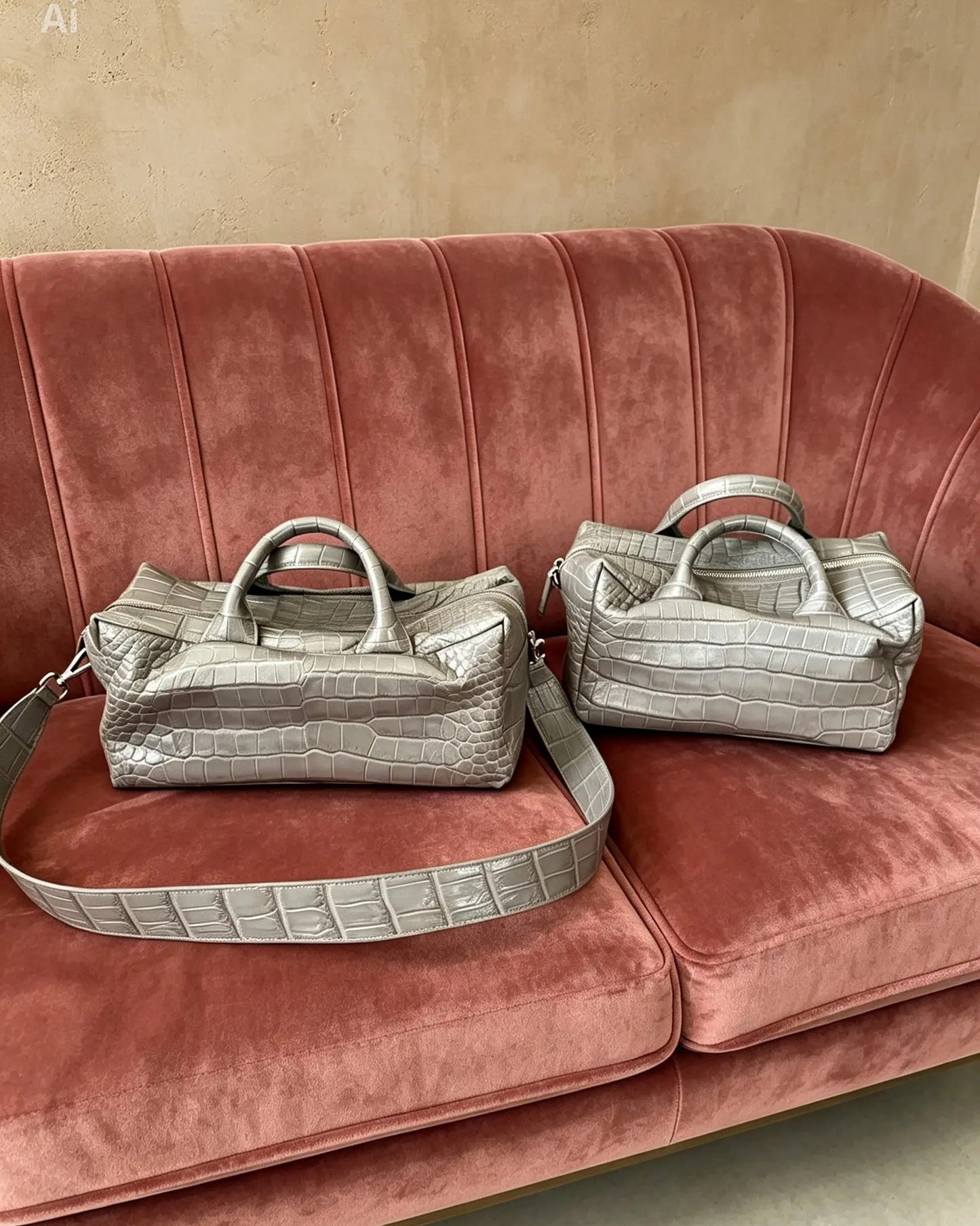 Two metallic silver crocodile-embossed handbags with top handles and detachable straps, displayed on a dusty rose velvet sofa against a beige wall.