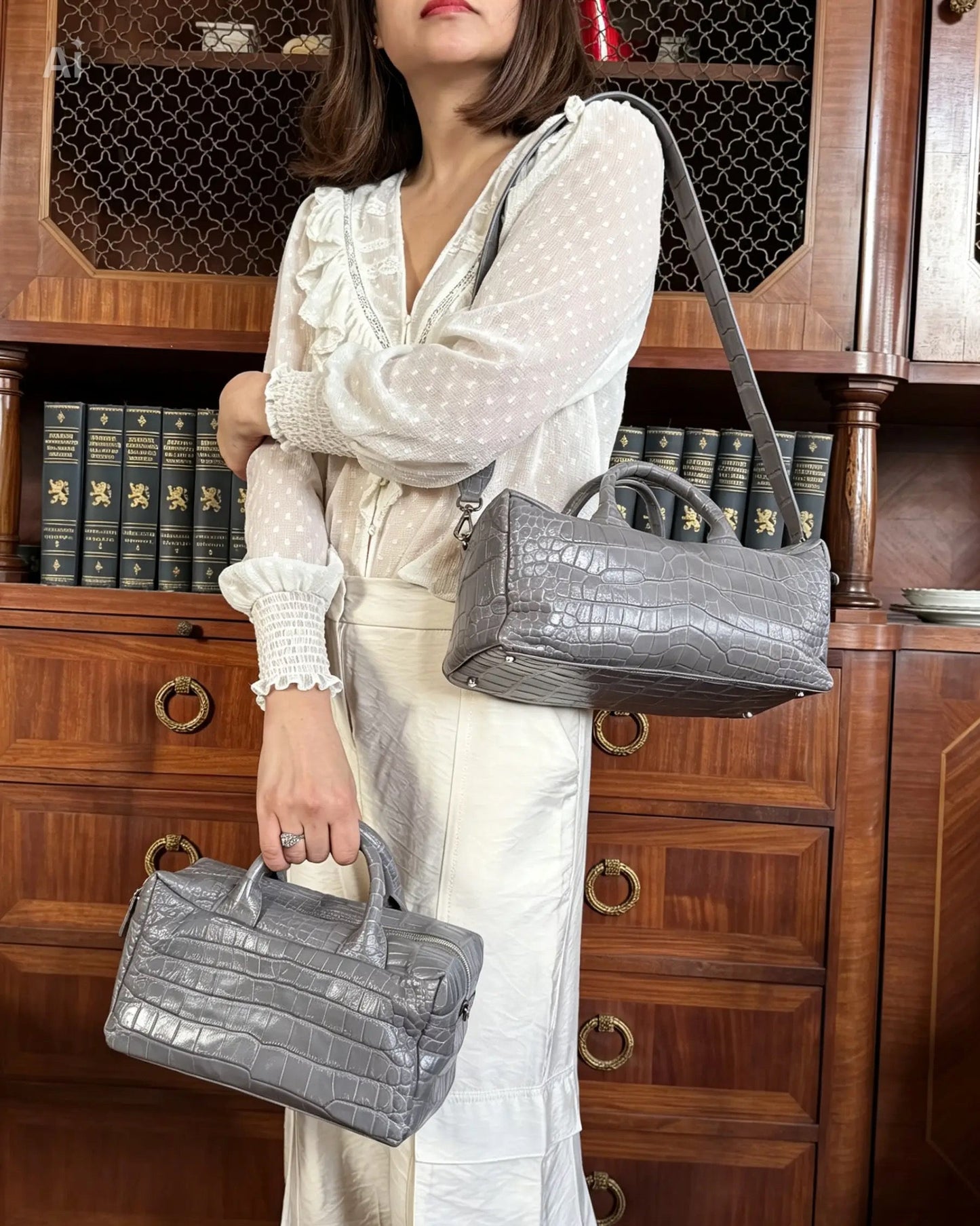 A woman wearing a white long-sleeve blouse and cream skirt holds two gray crocodile-embossed leather handbags in front of a dark wood bookcase.