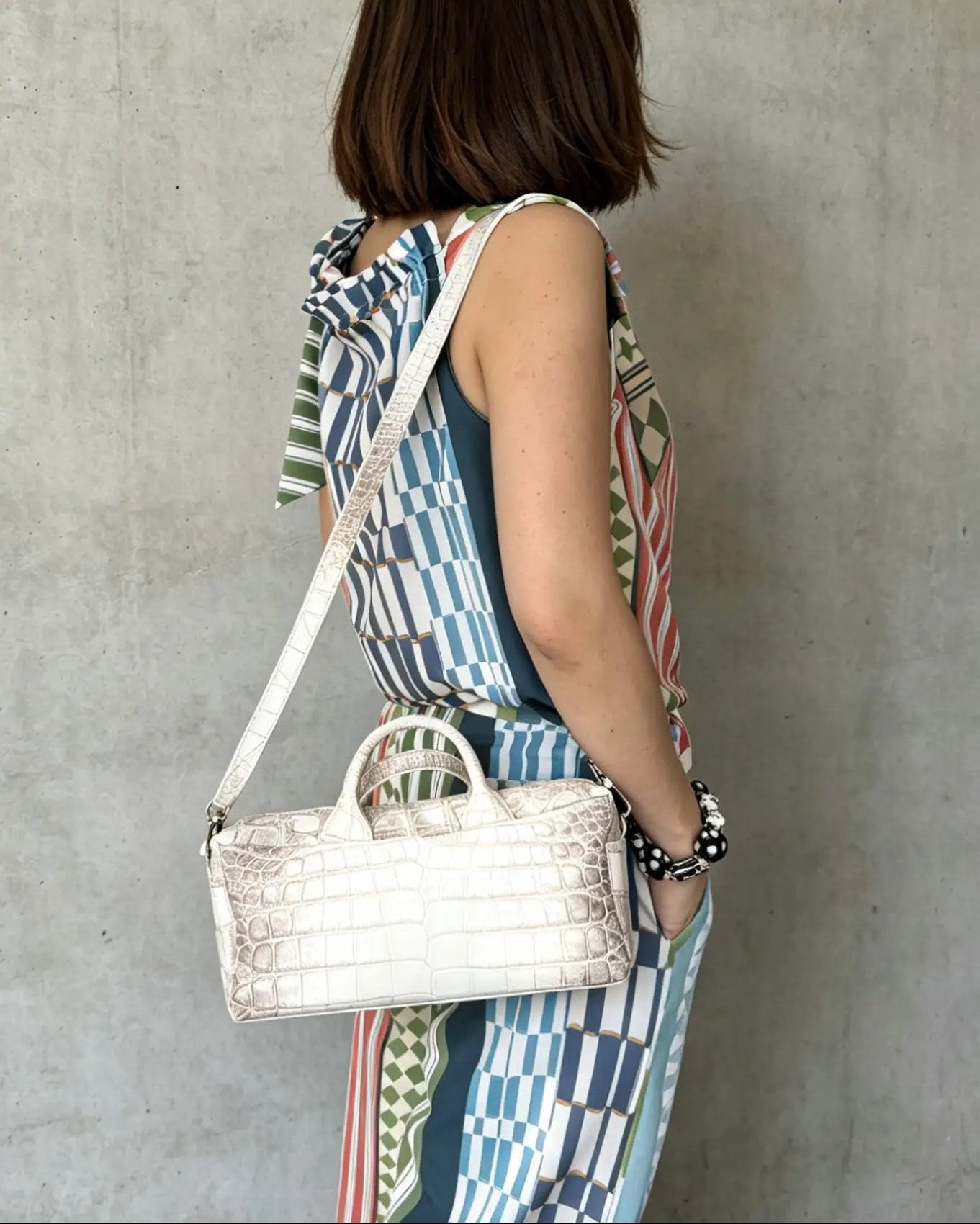 A woman with short brown hair is shown from behind, wearing a colorful geometric print dress and carrying a white crocodile-embossed handbag with a white shoulder strap against a concrete wall.
