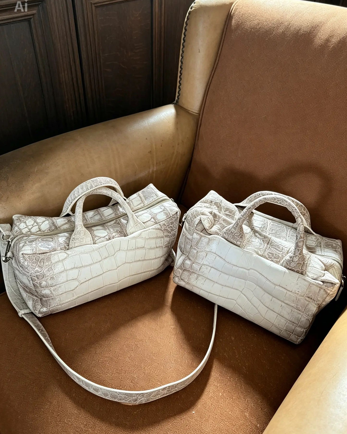Two small structured handbags with top handles and long shoulder straps, shown resting on a tan leather armchair. The bags have a glossy embossed crocodile or alligator texture in a light champagne or off-white tone.