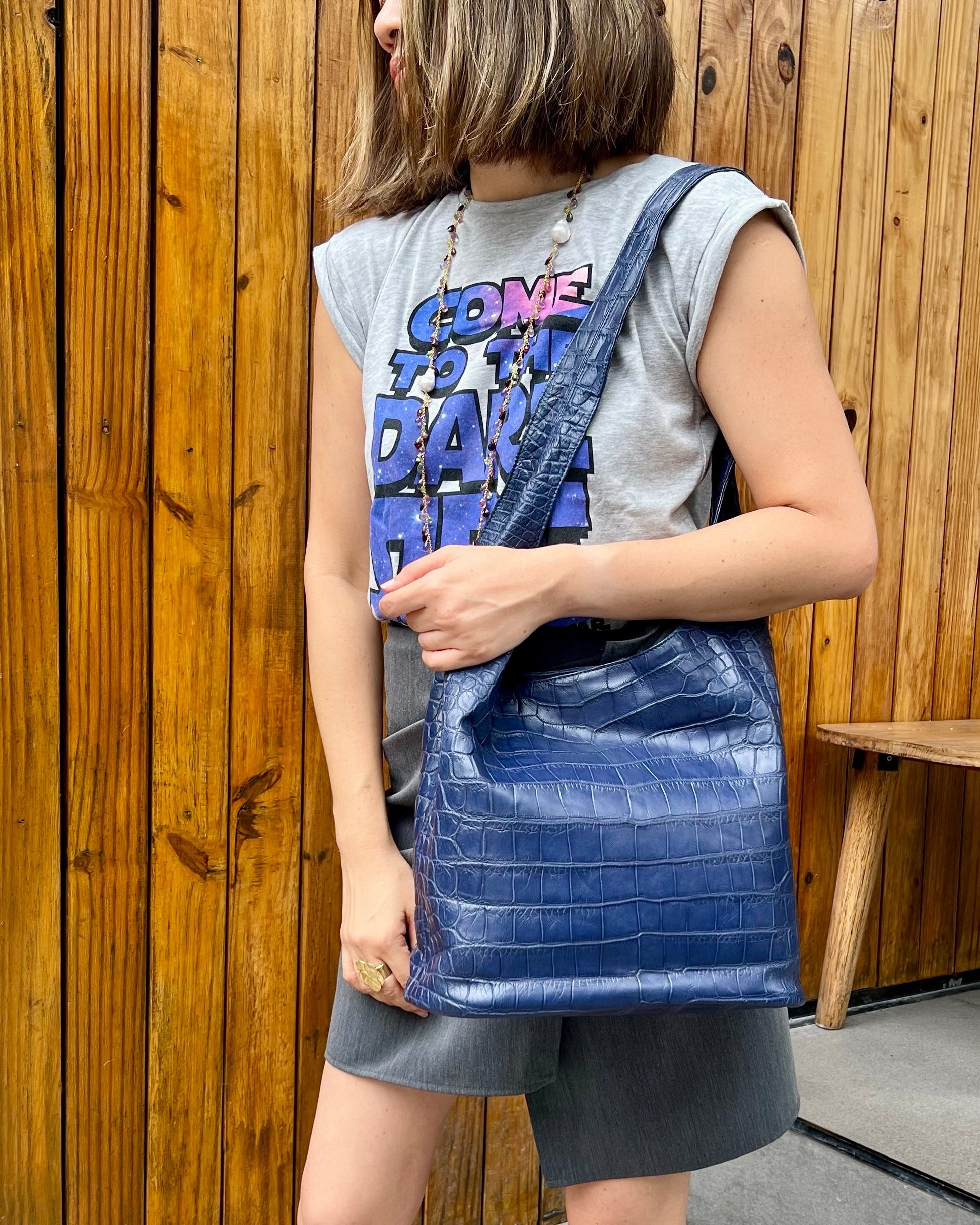 Woman holding a navy blue crocodile bag against a wooden wall.