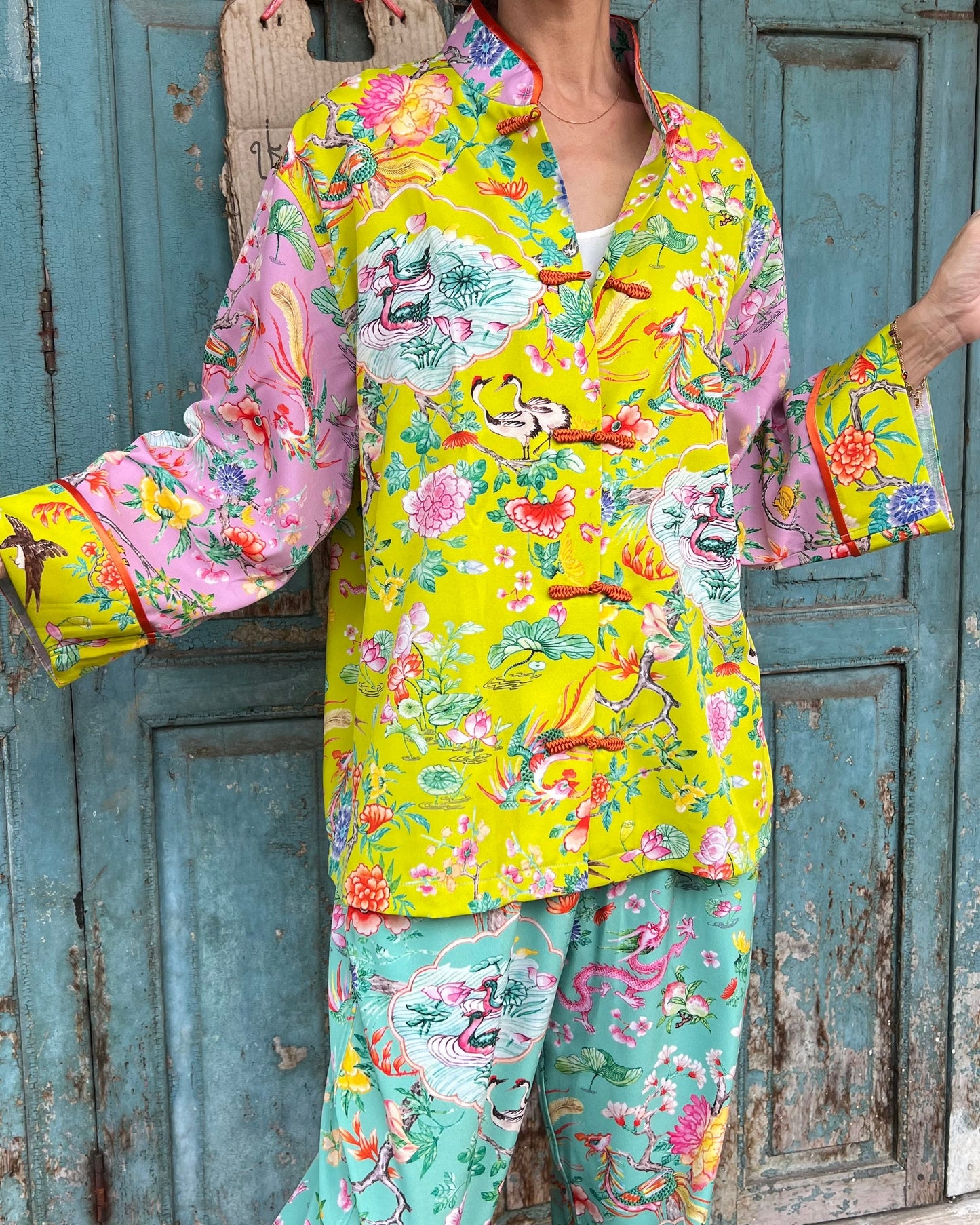 A woman wearing a lime-pink mandarin top with a floral pattern and Chinese-inspired details, paired with matching pants, standing in front of a blue door.