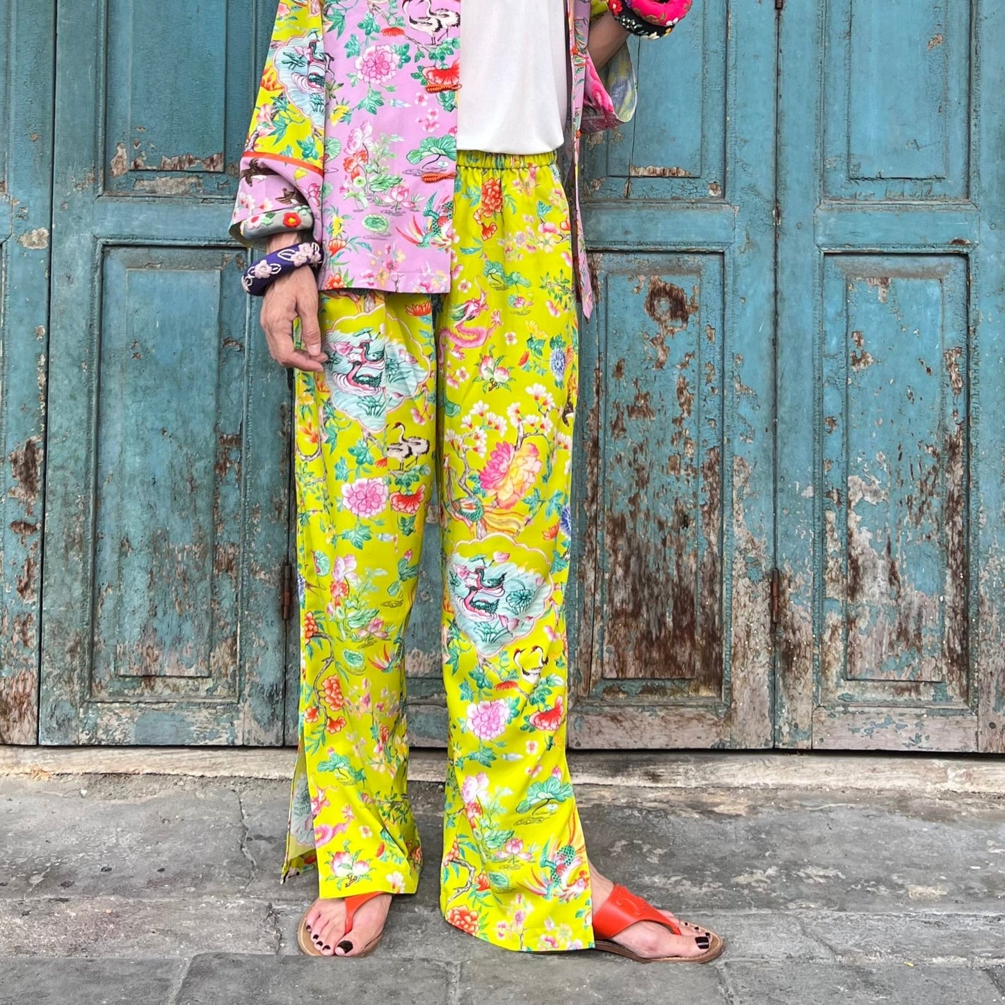 A person standing in front of a blue door wearing lime green pants with hand-drawn prints and side slits, paired with a white top and red sandals.