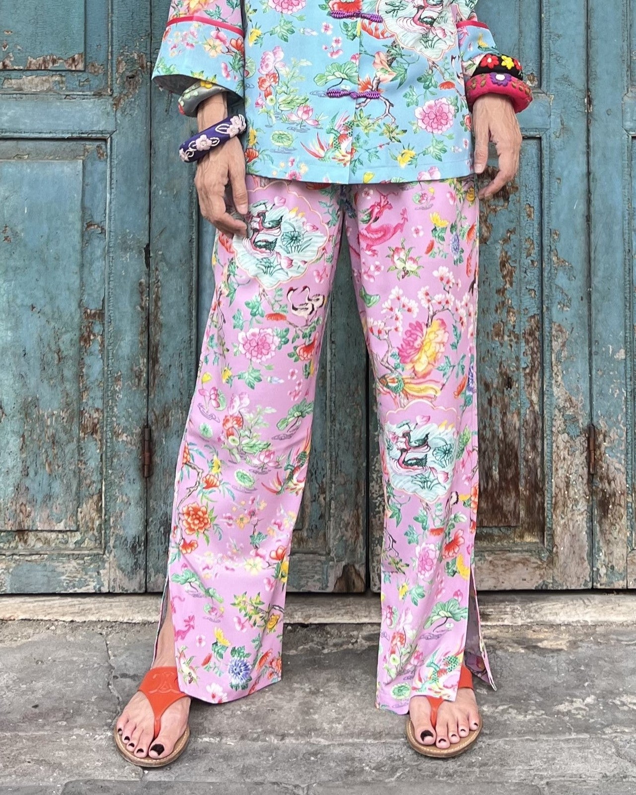 Person wearing a colorful floral outfit with pink pants and a patterned top against a rustic wooden door.