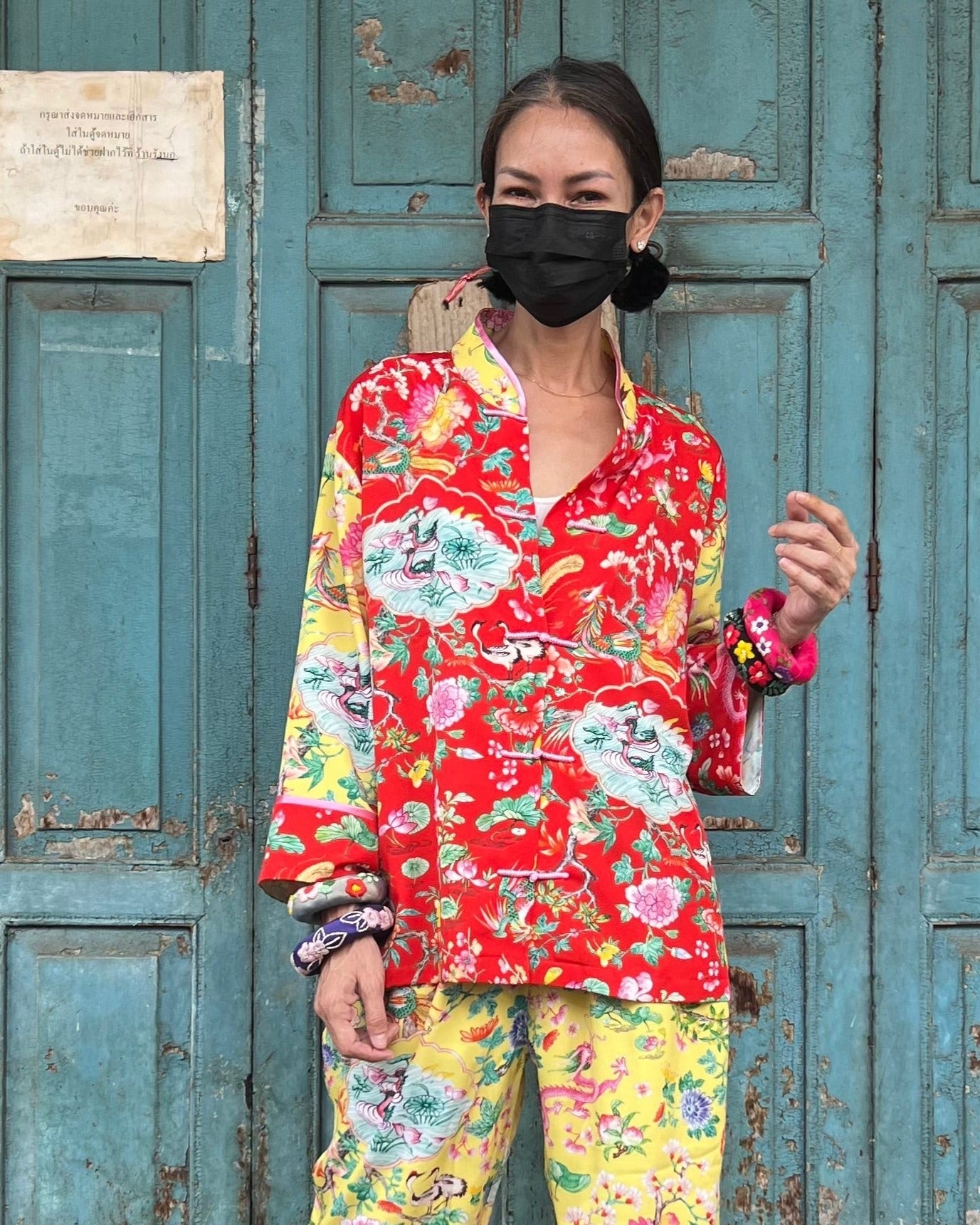 A woman wearing a red and yellow mandarin top with floral patterns and Chinese characters, paired with matching pants, standing in front of a blue door.