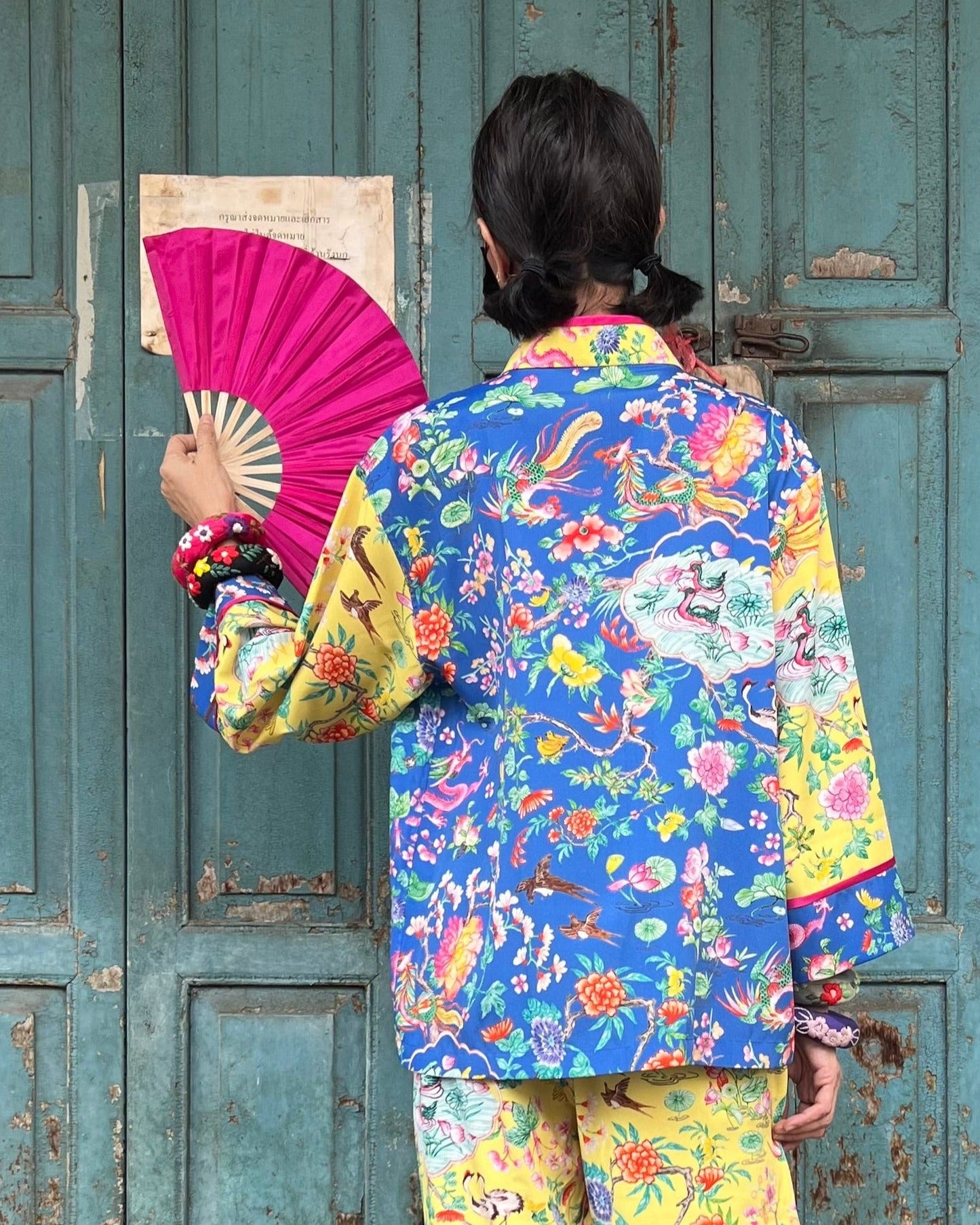 Person wearing a colorful traditional outfit with a pink fan against a rustic wooden door.