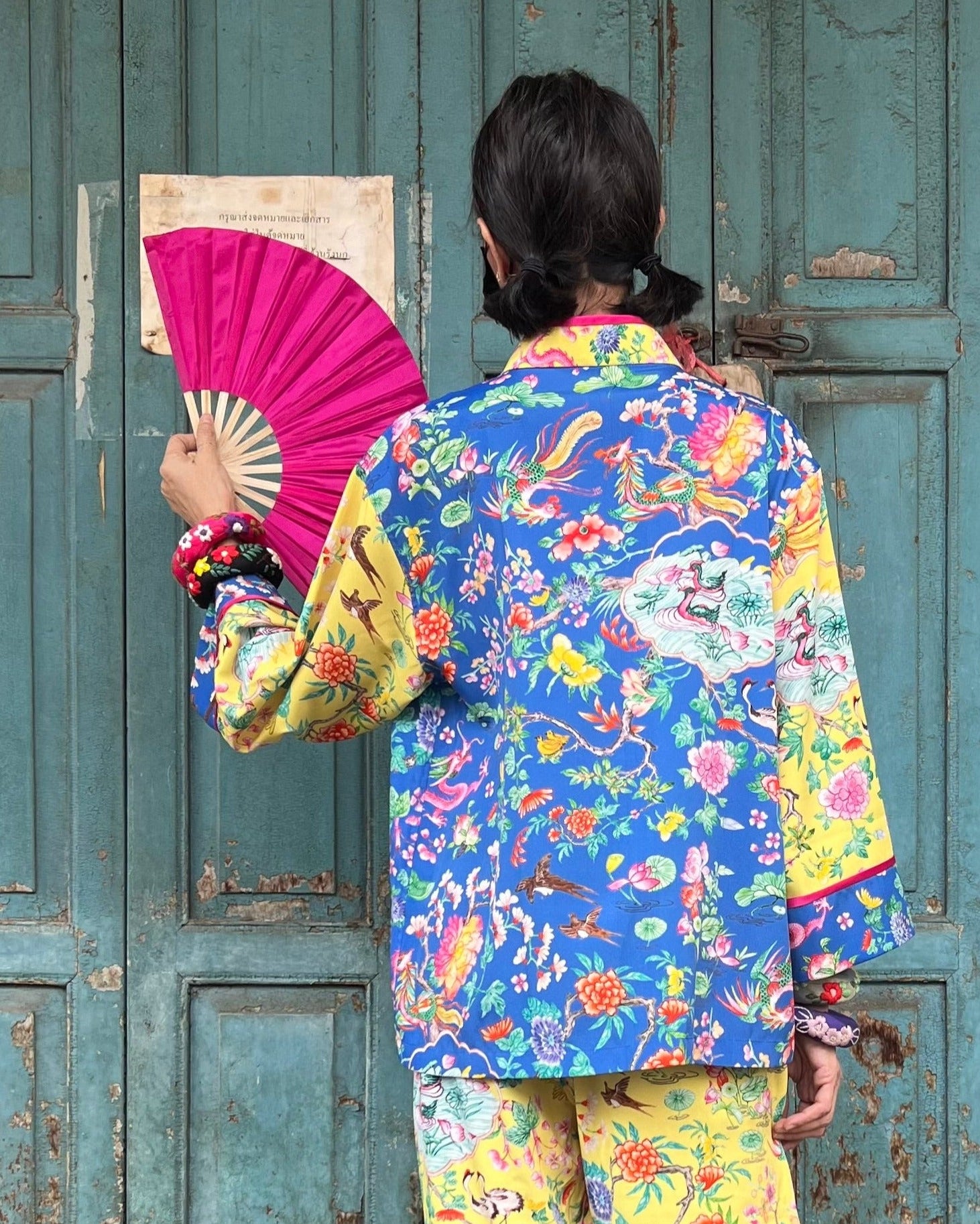 Person wearing a colorful traditional outfit with a pink fan against a rustic wooden door.