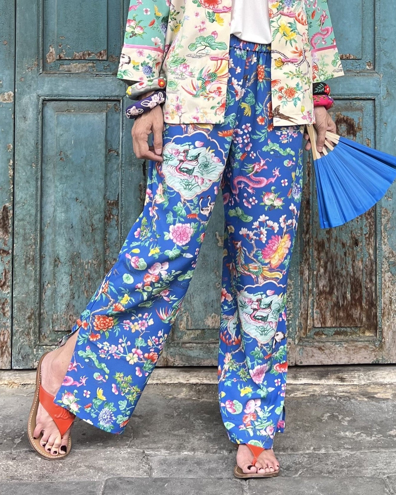 Person wearing colorful floral pants and jacket with a blue fan, standing in front of a rustic wooden door.