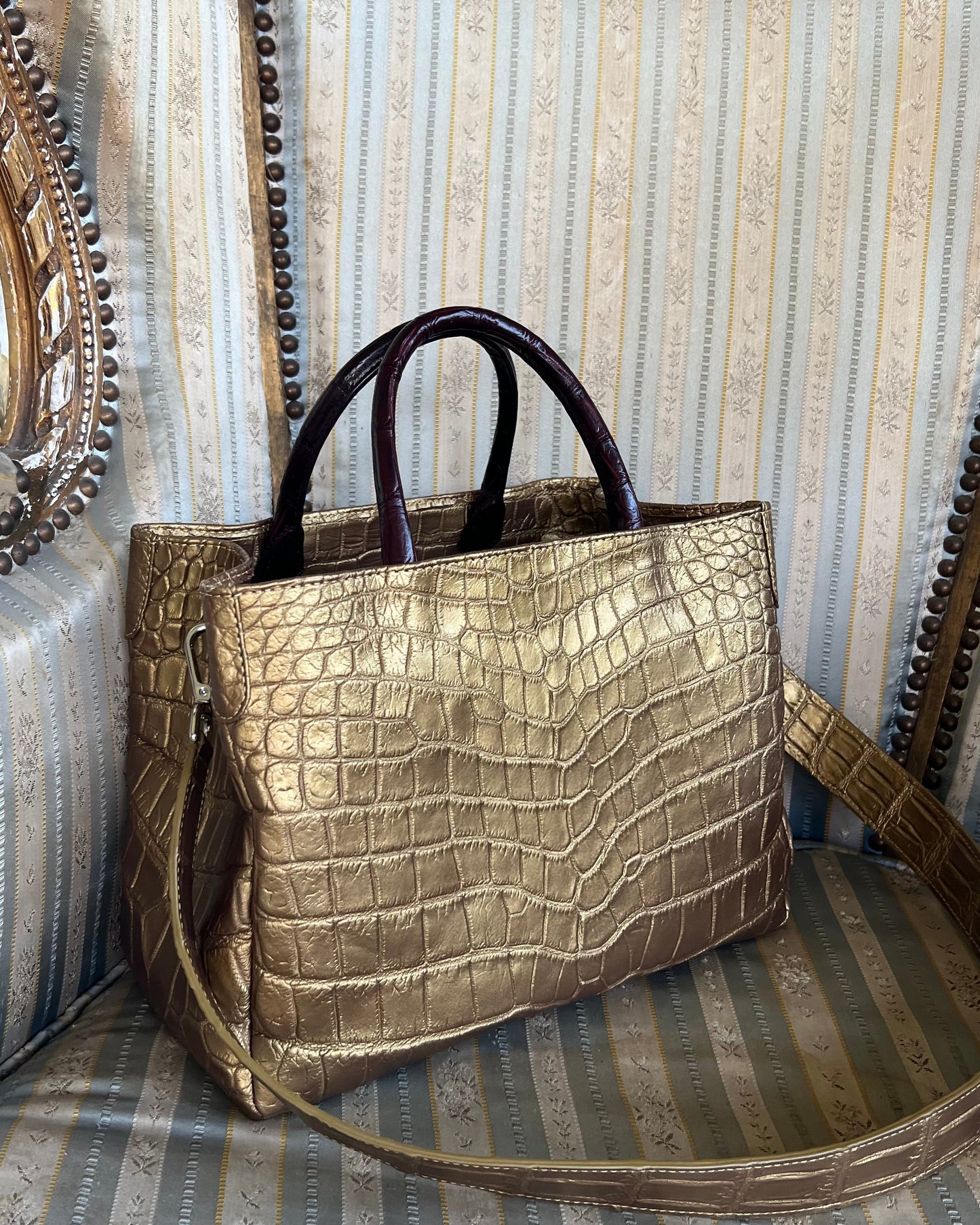 A gold-colored, crocodile-patterned leather handbag with a beige lining, placed on a striped chair.