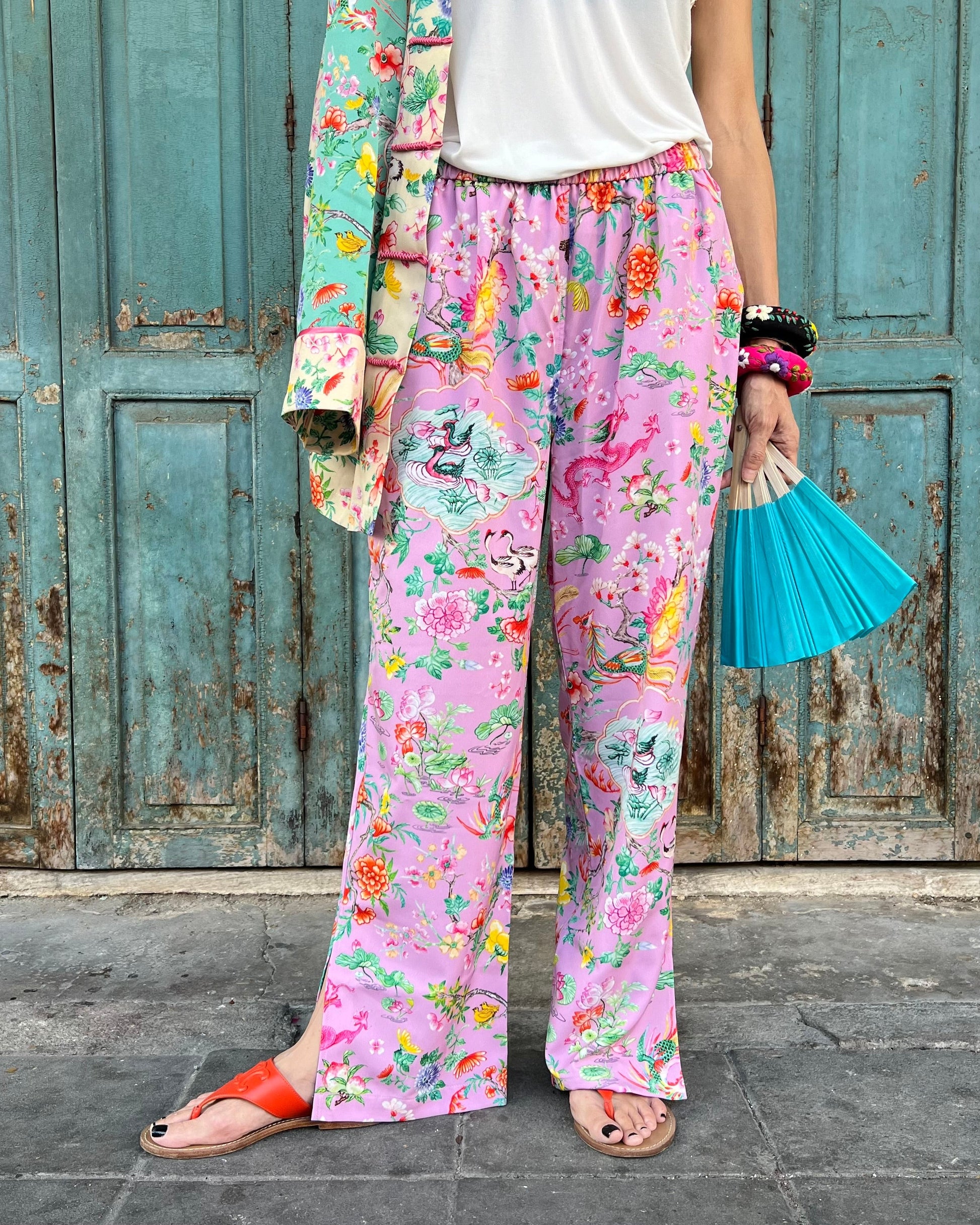 Person wearing colorful floral pants and a jacket, holding a blue fan, in front of a rustic wooden door.
