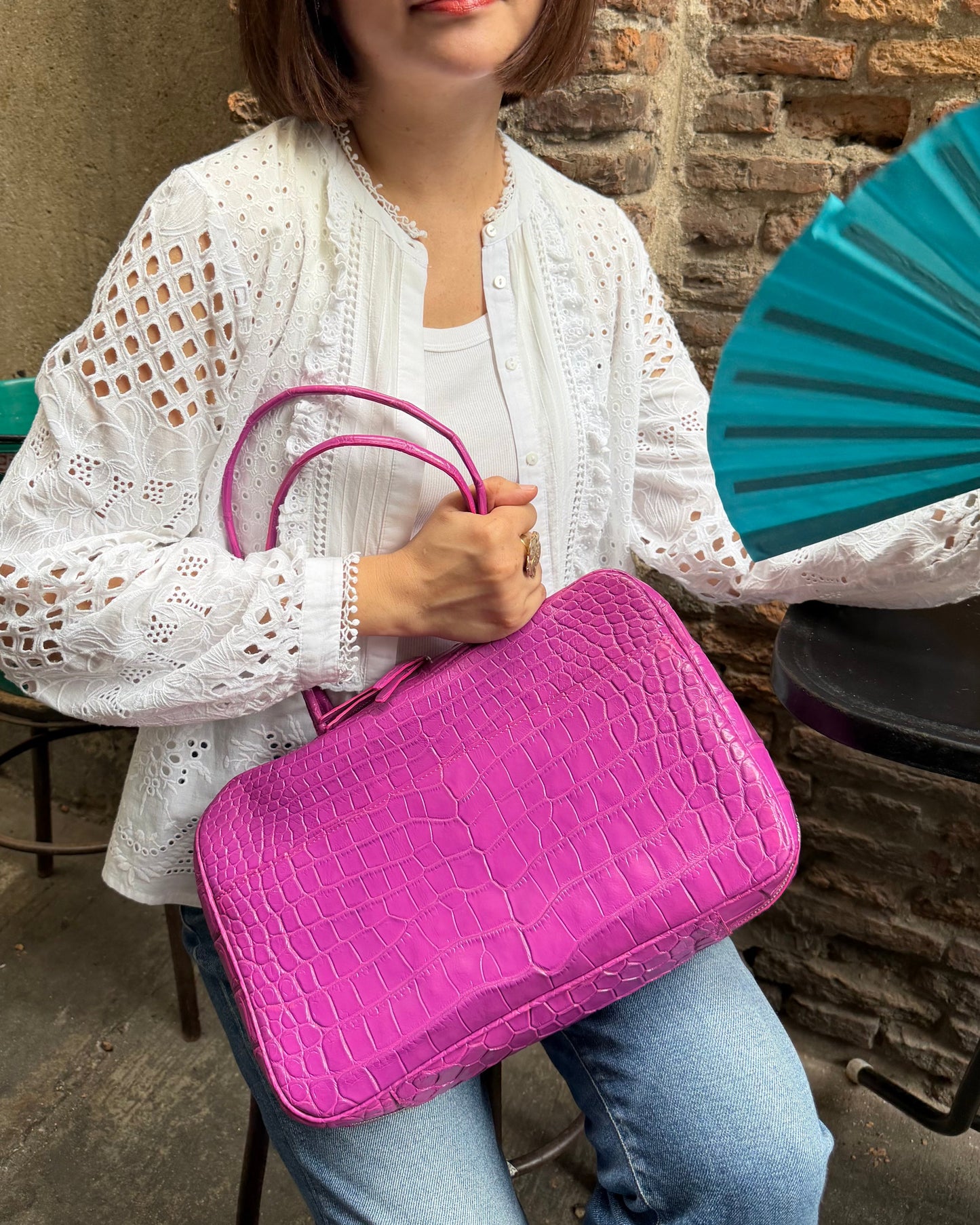 A woman wearing a white eyelet cardigan and light blue jeans holds a bright pink crocodile-embossed handbag with double top handles in a rustic indoor setting with a brick wall and teal folding fan.