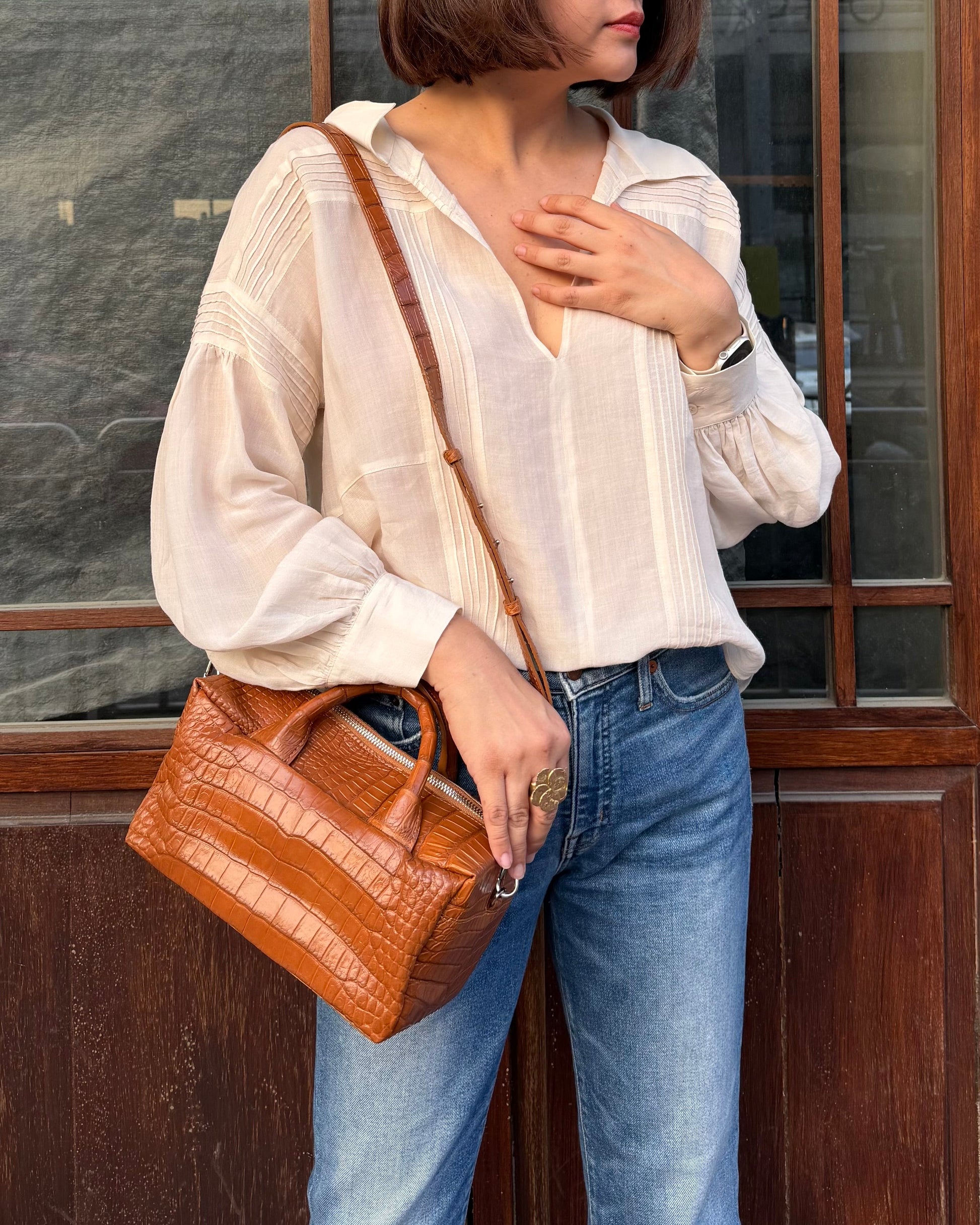 Person holding a brown handbag wearing a white shirt and blue jeans, standing against a wooden door.