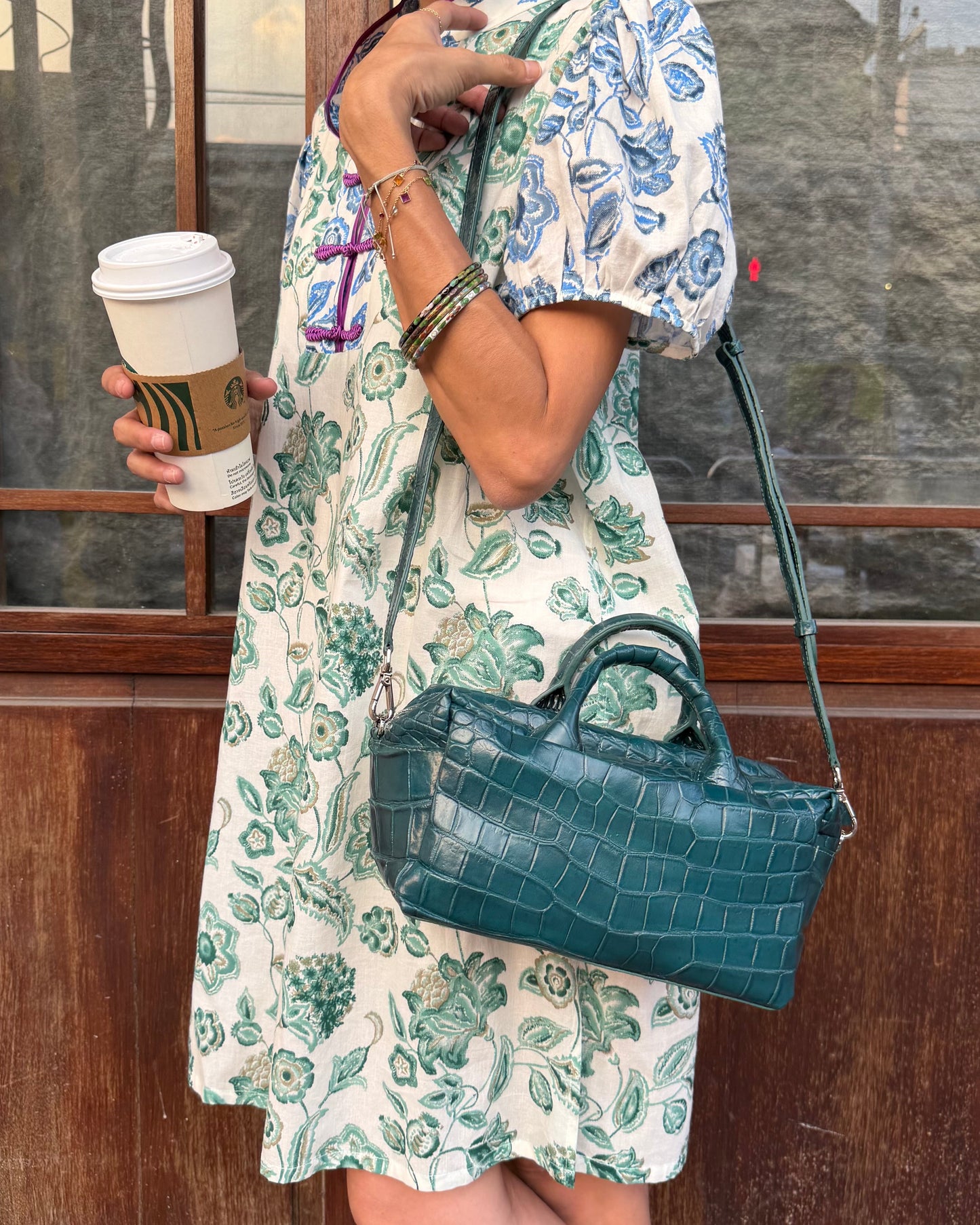 Woman in a floral dress holding a coffee cup and a green handbag against a wooden wall.