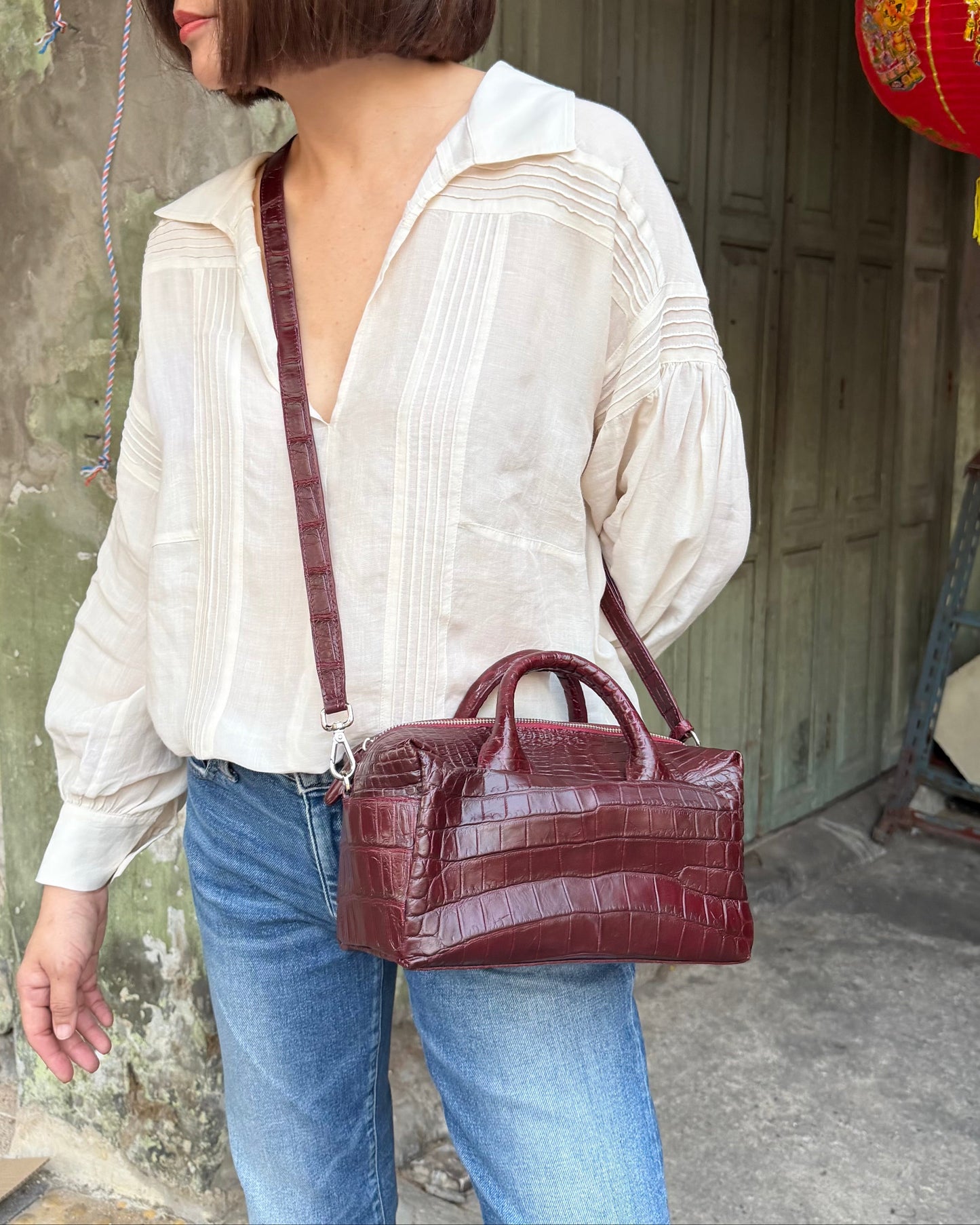 Person wearing a white blouse and blue jeans with a brown handbag, standing in front of a rustic building with a red lantern.