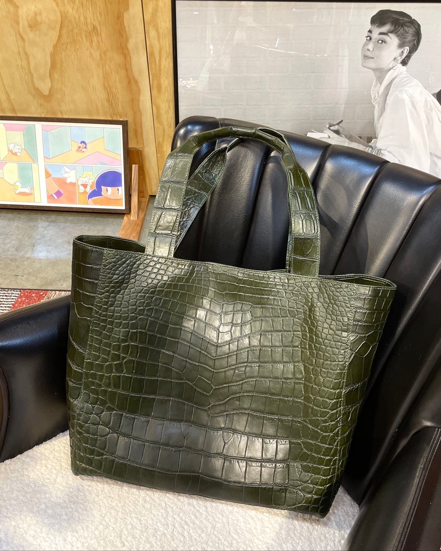 Olive green textured tote bag on a black leather couch with framed pictures in the background.