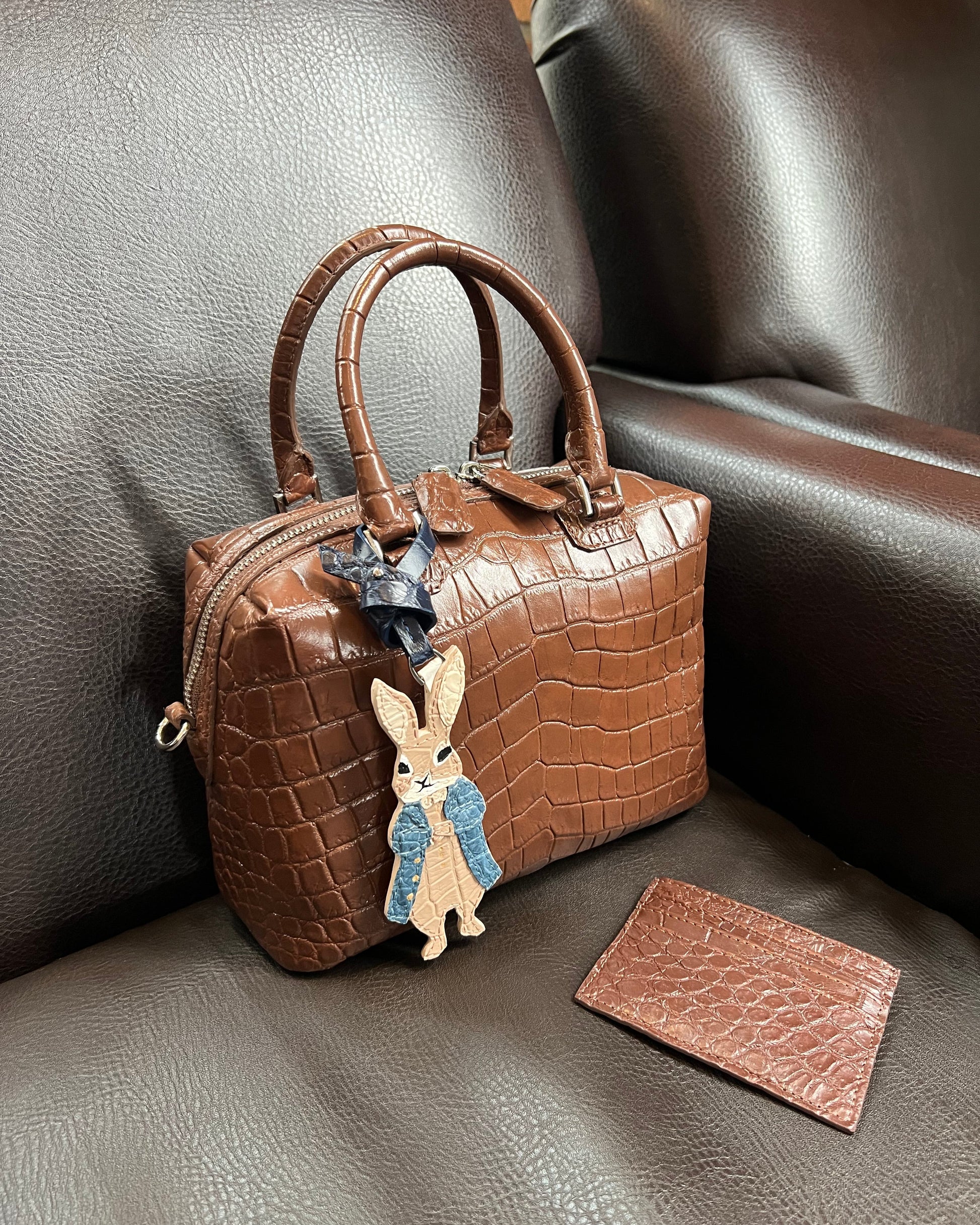A brown crocodile-embossed leather handbag with double top handles sits on a dark brown leather armchair. A matching small leather card holder lies beside it, and the bag is decorated with a Peter Rabbit character charm.