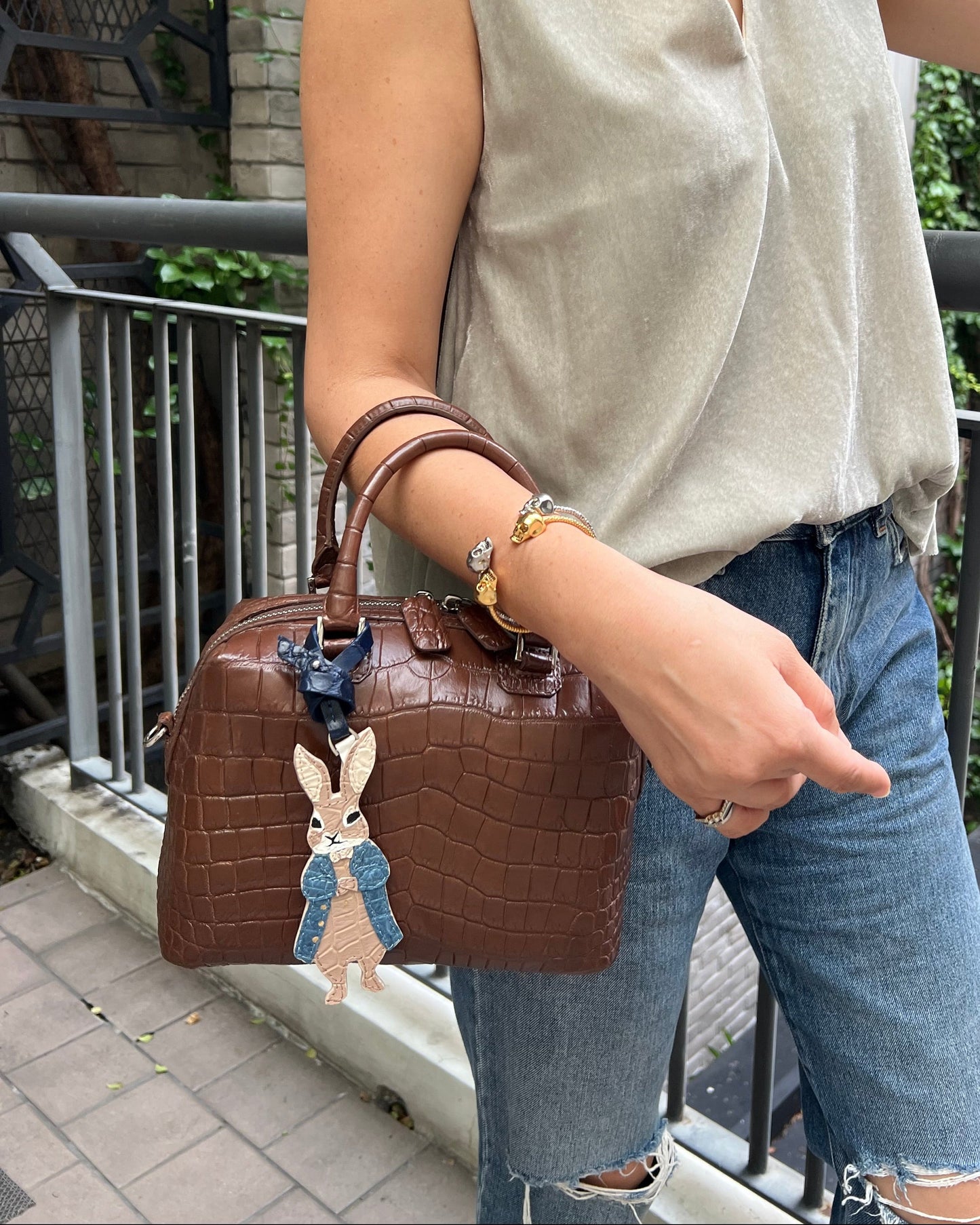 A woman wearing a beige sleeveless top and blue jeans carries a brown crocodile-embossed handbag with a rabbit charm in an outdoor setting.