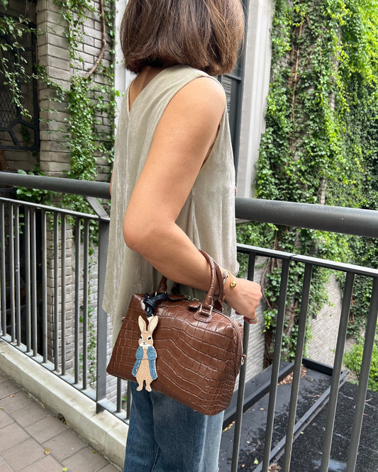 A woman wearing a beige sleeveless top and blue jeans carries a brown crocodile-embossed handbag with a rabbit charm on a balcony with green ivy.