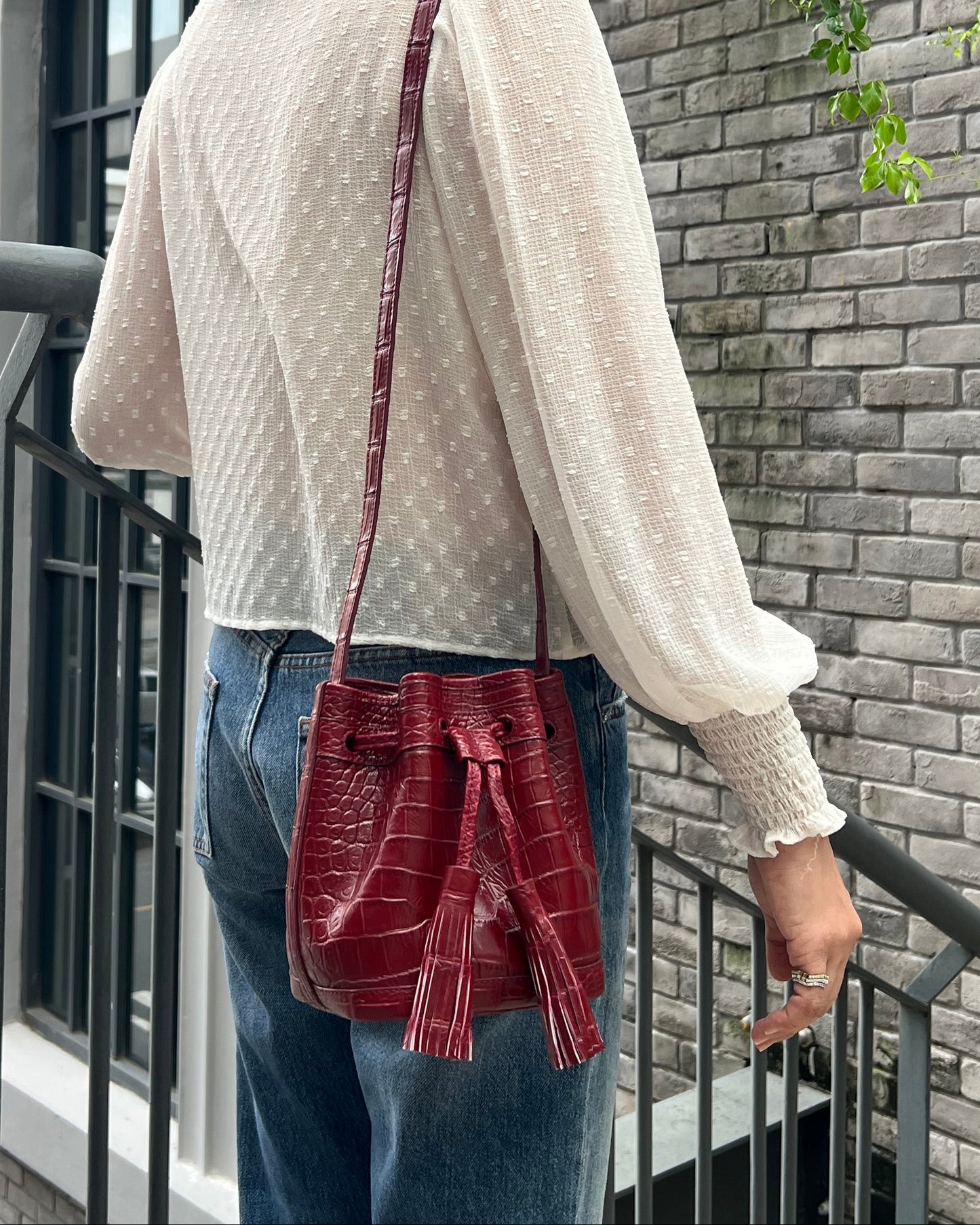 A person wearing a cream-colored textured knit sweater, blue jeans, and a red quilted crossbody bag, photographed from a high-angle perspective against a gray brick building background.