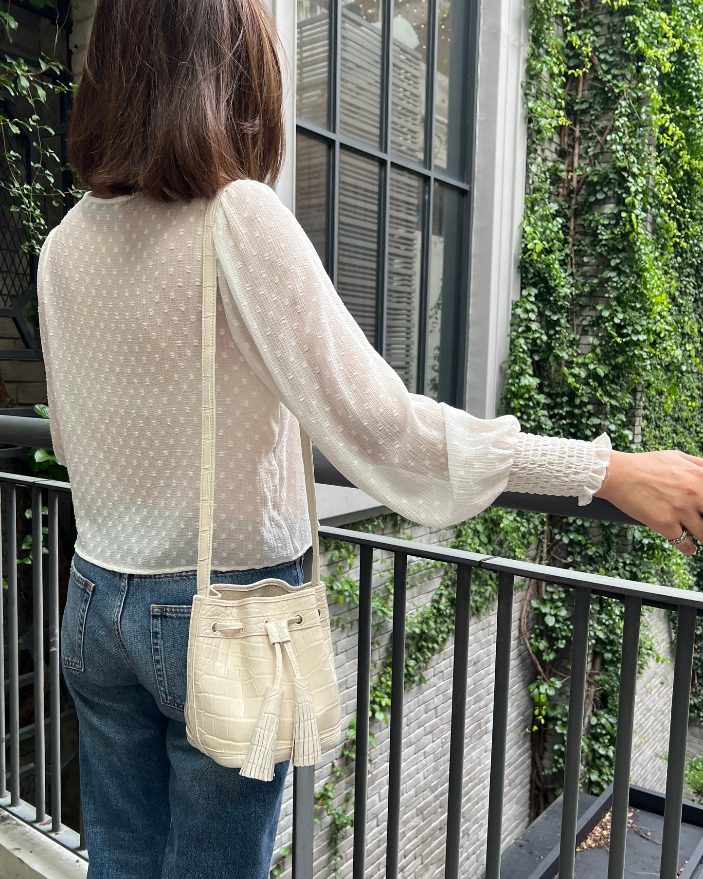 A woman wearing a white long-sleeve polka-dot blouse, blue jeans, and a cream crossbody bag, standing on a balcony with a green ivy-covered wall in the background.
