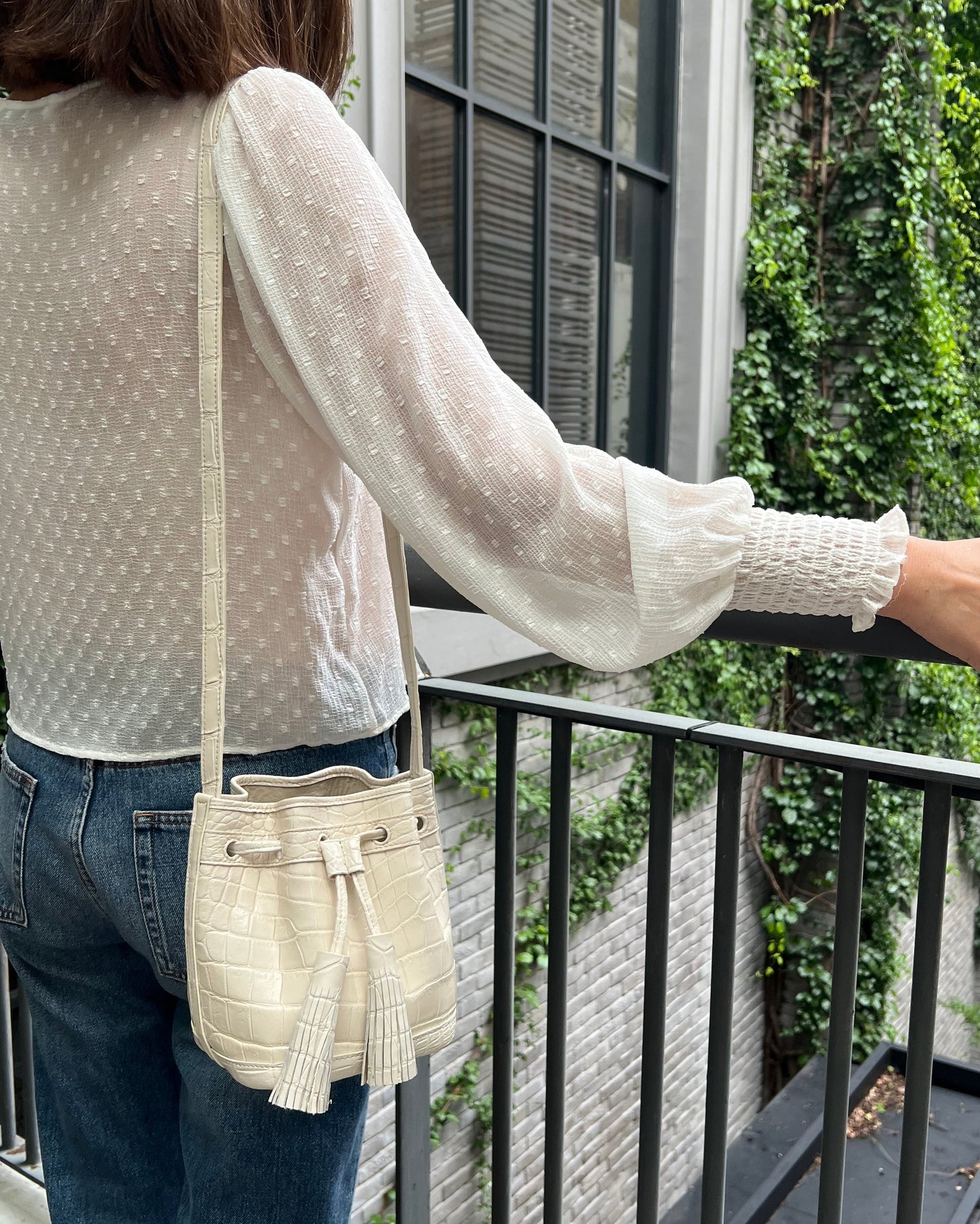 A woman wearing a sheer white polka-dot blouse and blue jeans stands on a balcony, carrying a cream crocodile-embossed bucket bag with a tassel strap.