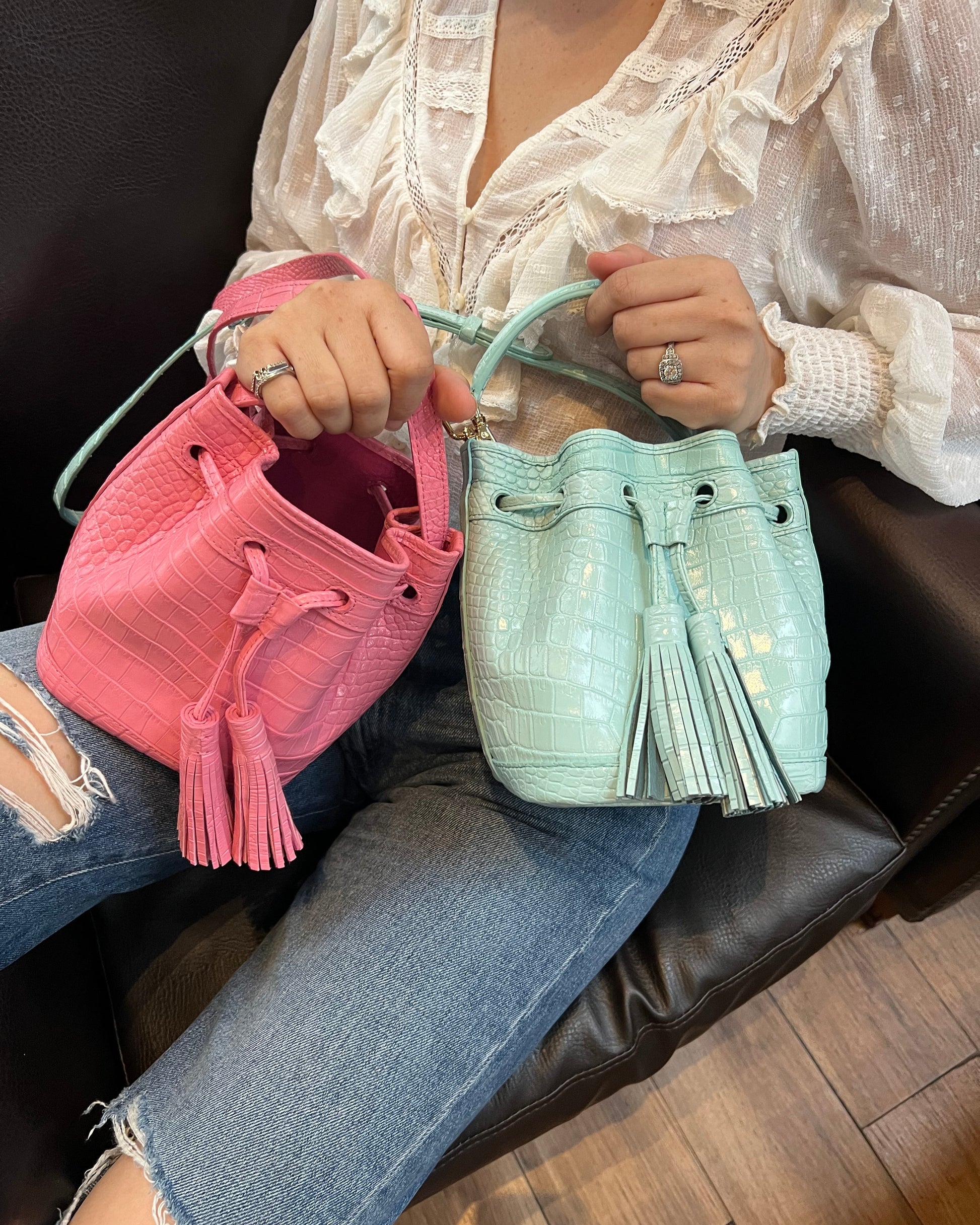 A person seated on a dark leather chair holds two bucket-style drawstring handbags, one in light mint green and one in pink, with blue ripped jeans and a white blouse visible.