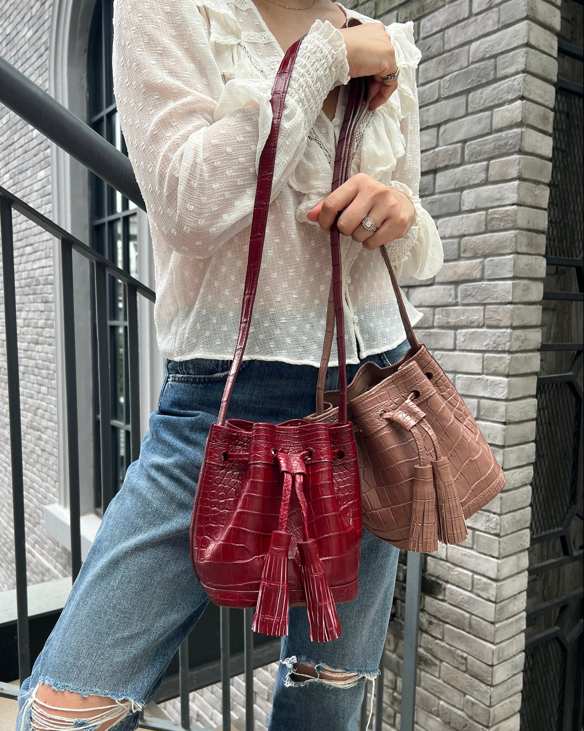 A person wearing a white textured blouse and ripped blue jeans holds up two shoulder bags. One bag is a dusty rose pink with a crocodile-embossed texture, and the other is a deep burgundy red with a matching crocodile-embossed pattern and tassel detail.
