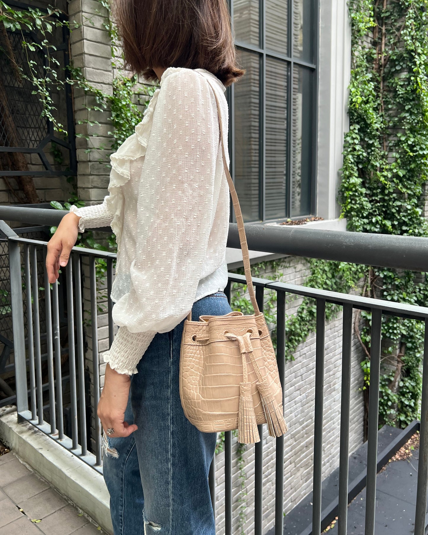 A woman wearing a white long-sleeve polka dot blouse and blue jeans, carrying a beige woven bucket bag with tassel details over her shoulder, standing on an outdoor balcony.