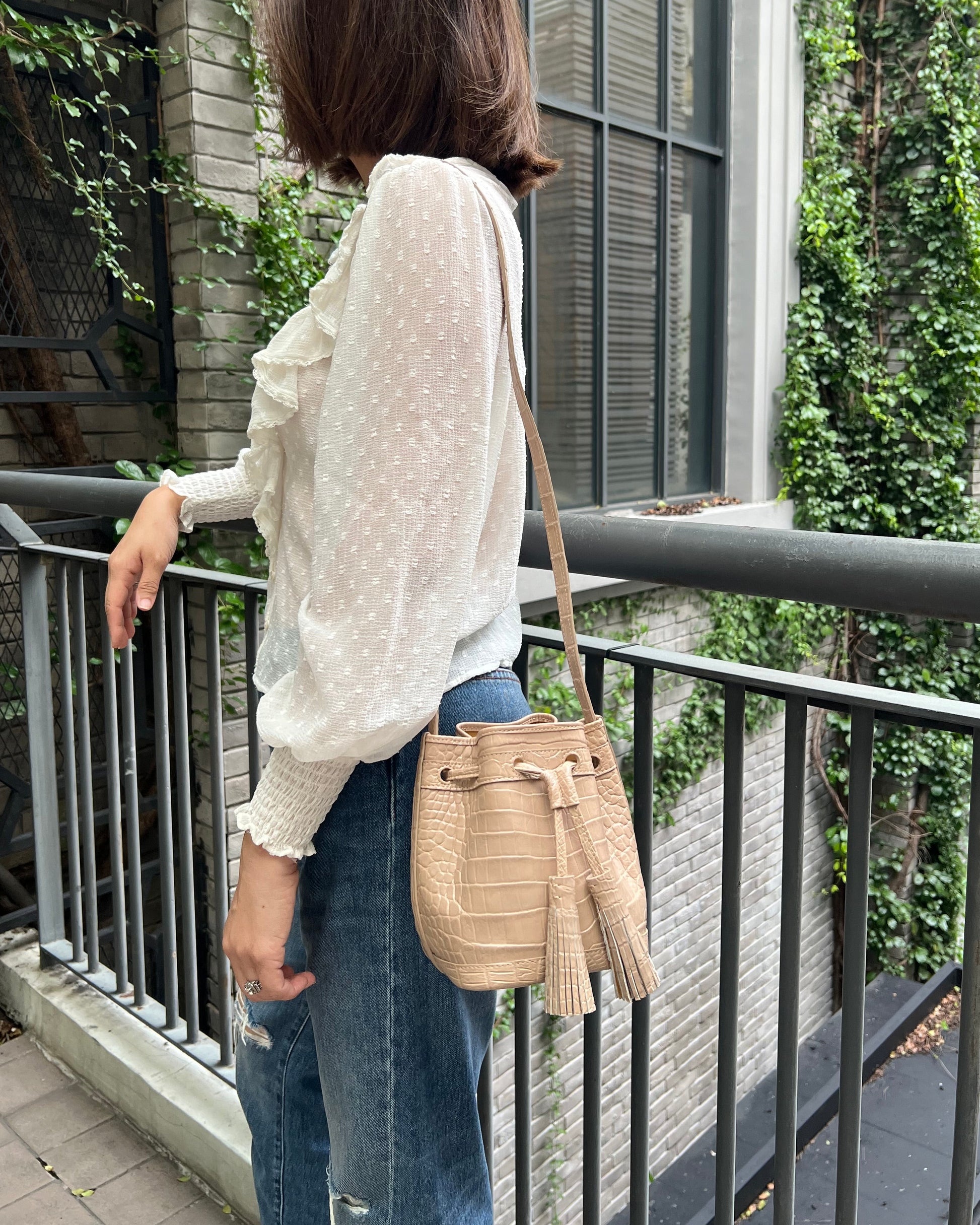 A woman wearing a white long-sleeve polka dot blouse and blue jeans, carrying a beige woven bucket bag with tassel details over her shoulder, standing on an outdoor balcony.