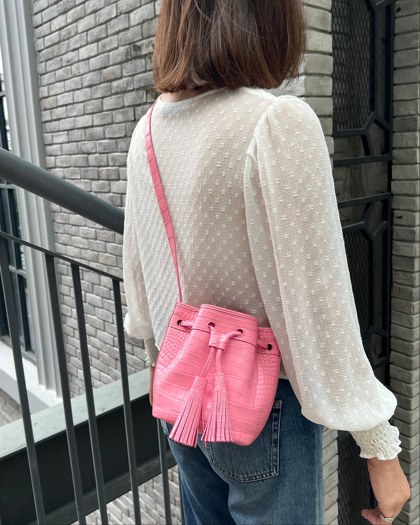 A woman viewed from behind wearing a white long-sleeve textured blouse and blue jeans, carrying a bright pink textured crossbody bag with a tassel drawstring against a gray brick wall.