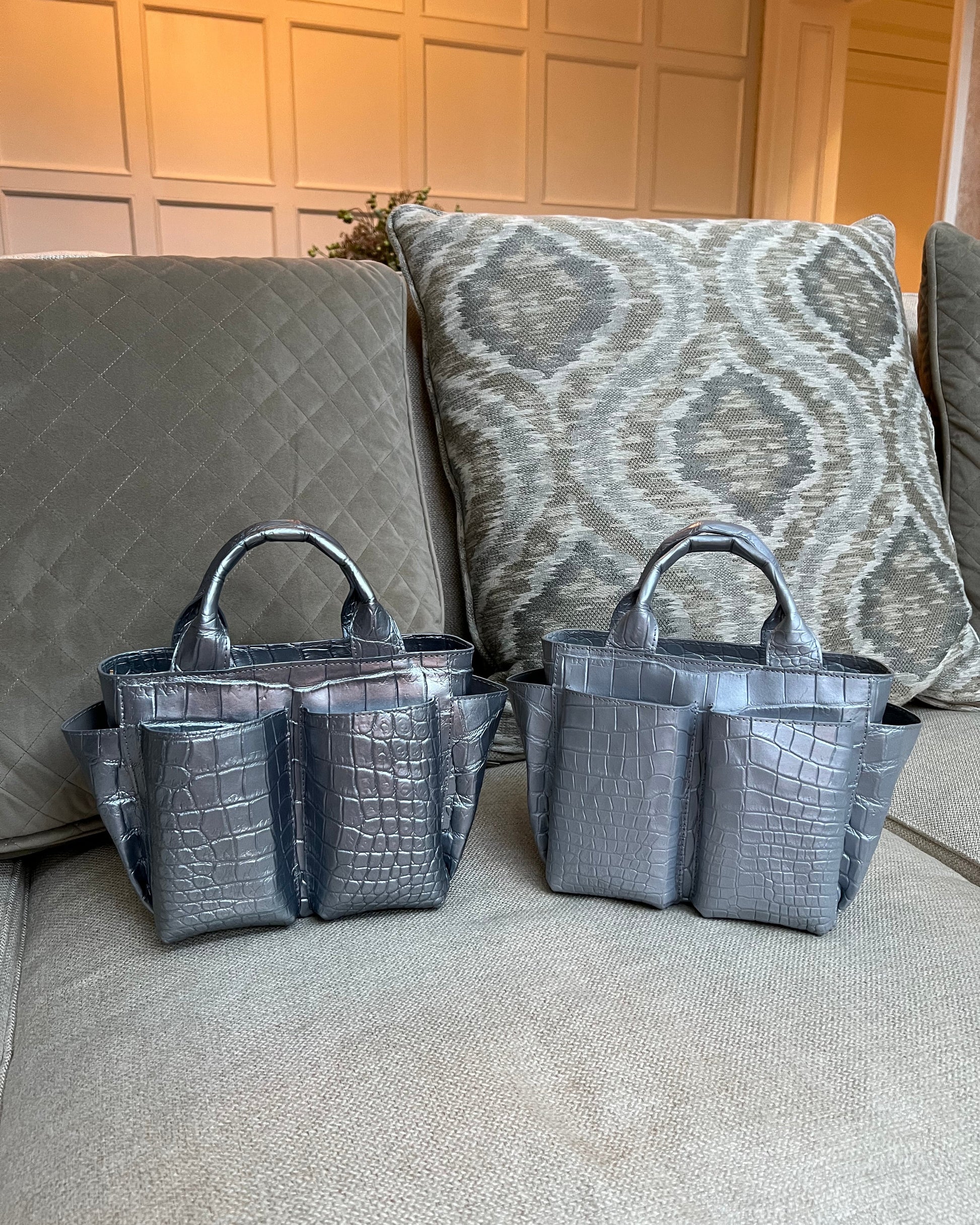 Two gray handbags on a textured surface with a patterned cushion in the background.
