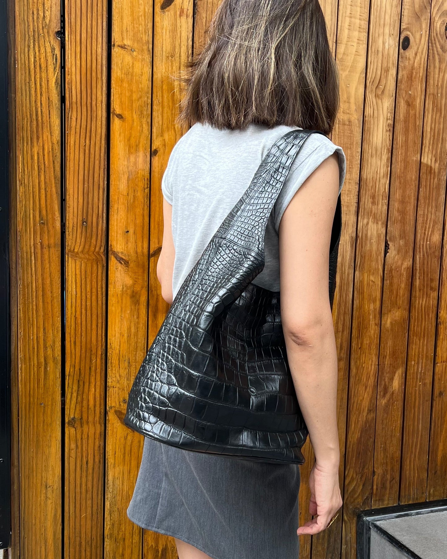 Person holding a black crocodile skin bag against a wooden wall.