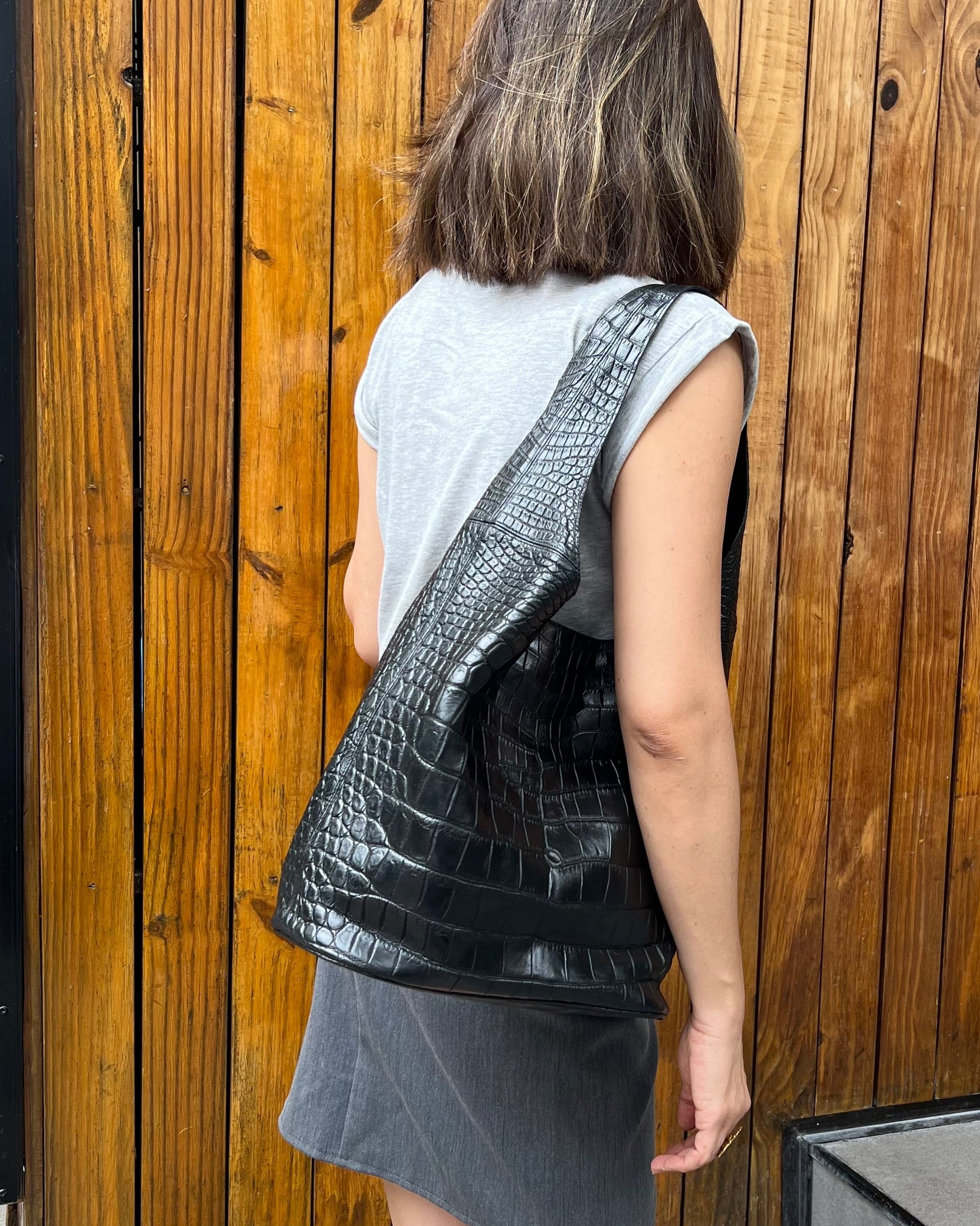 Person holding a black crocodile skin bag against a wooden wall.