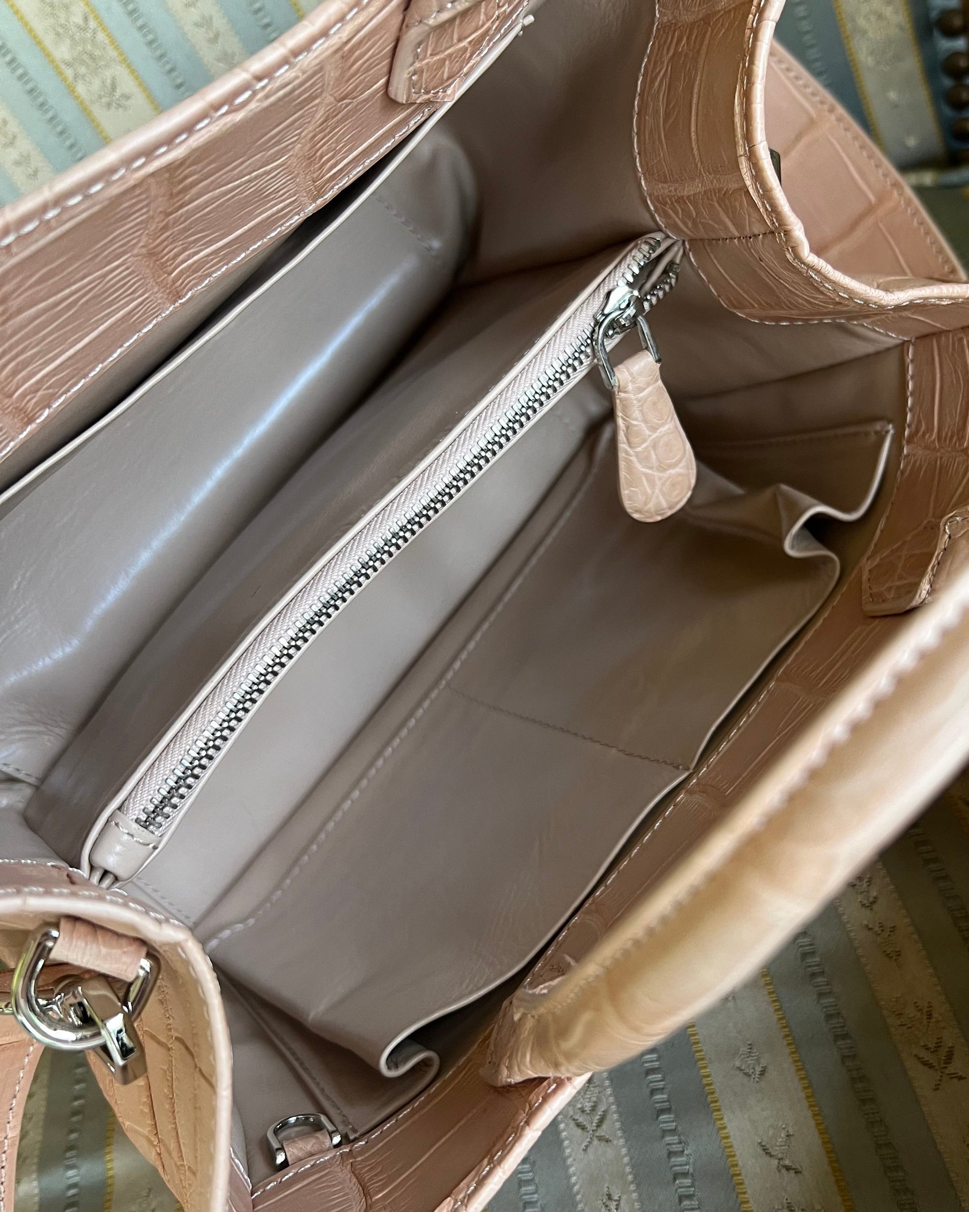 Close-up of the interior of a structured handbag in light beige faux crocodile leather, showing a zippered compartment and interior lining, set against a patterned fabric background.