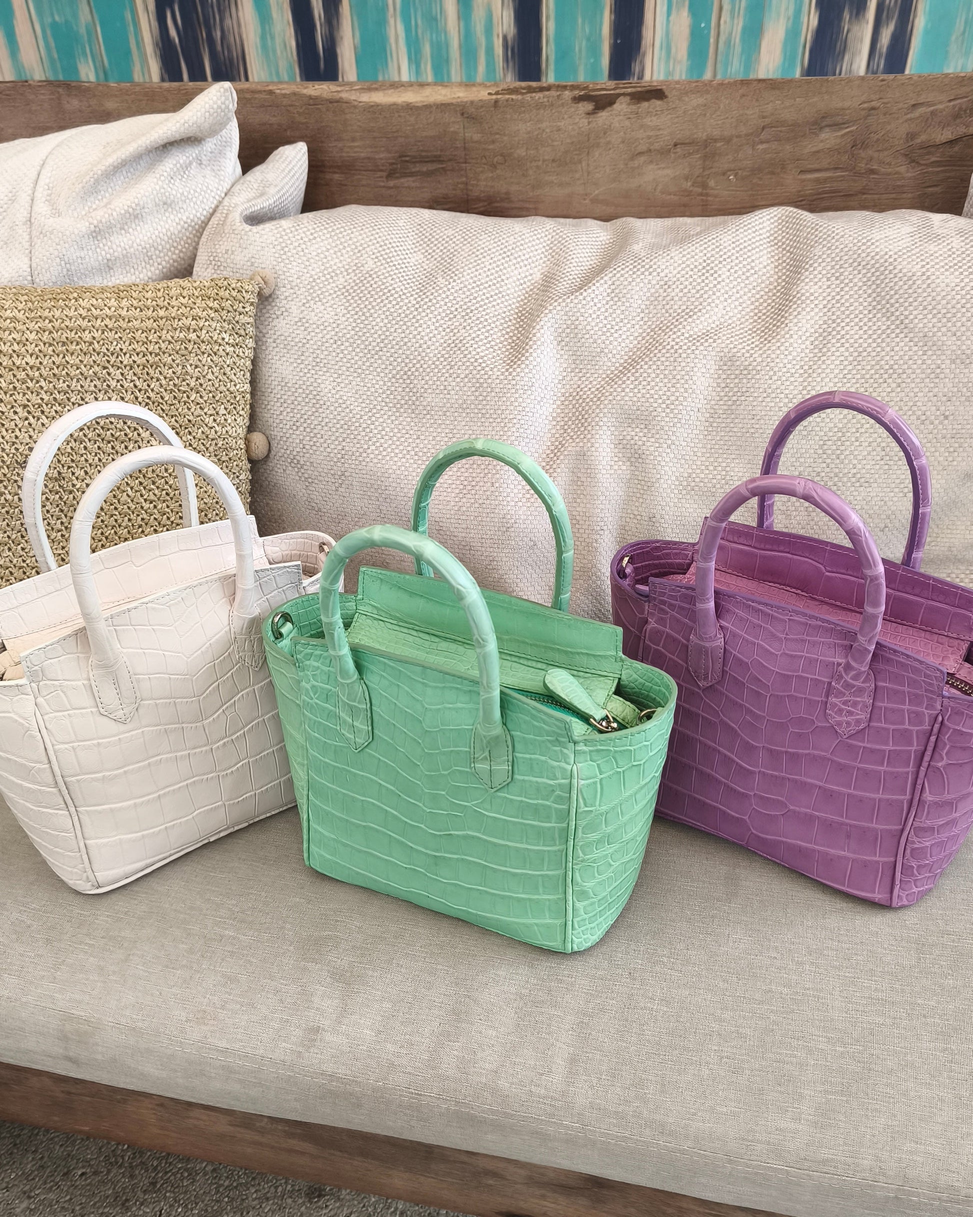 Three handbags in white, green, and purple on a couch with pillows in the background.