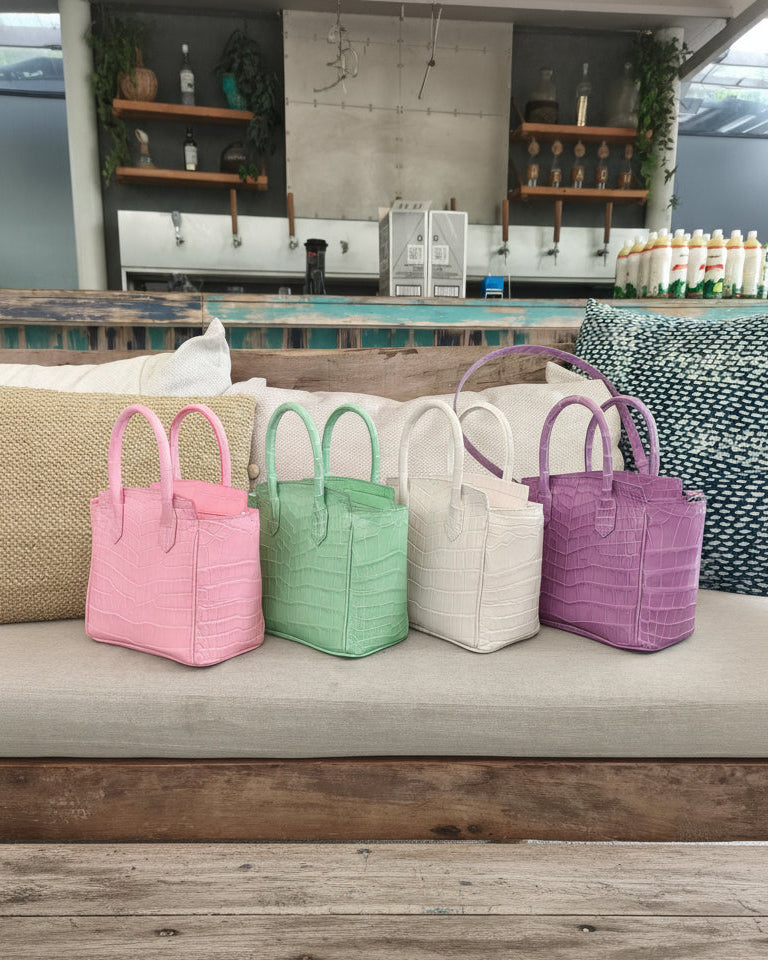 Four colorful handbags (pink, green, white, purple) on a couch with a coffee shop background.