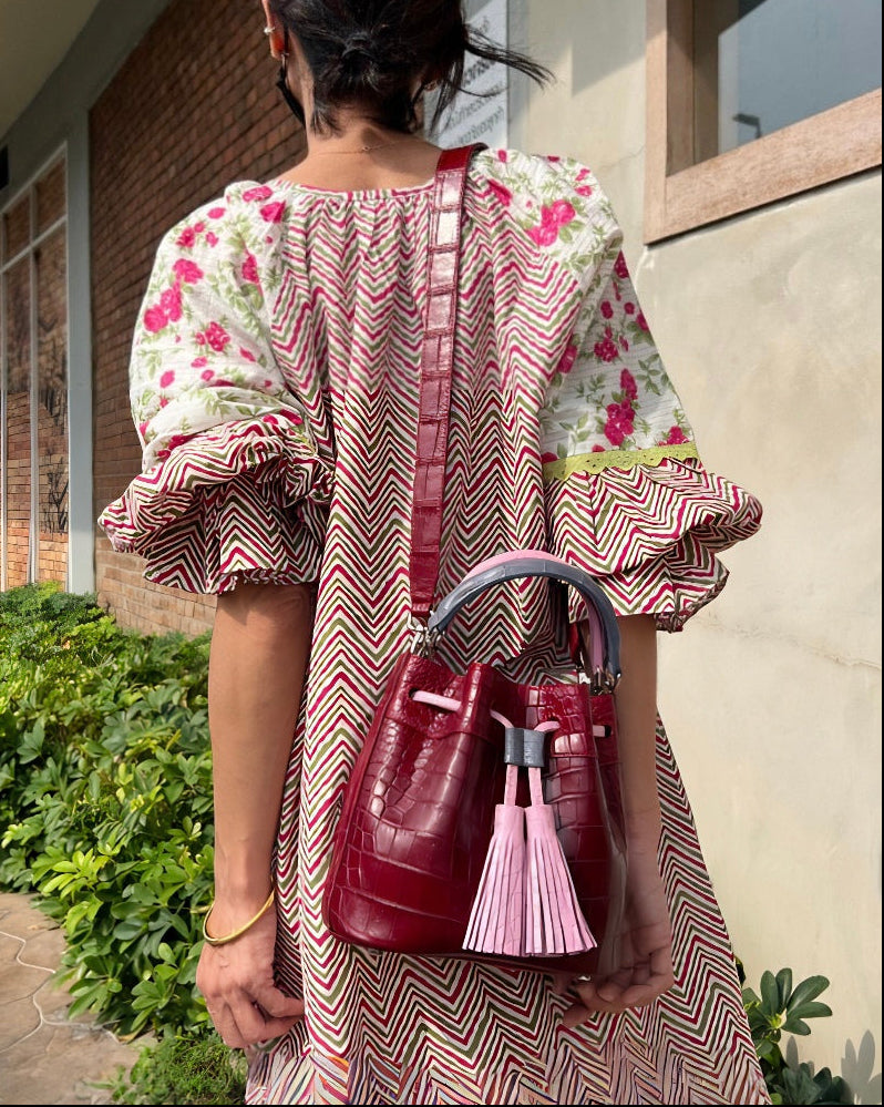 Woman in a floral dress holding a red handbag with a pink tassel, standing outdoors.