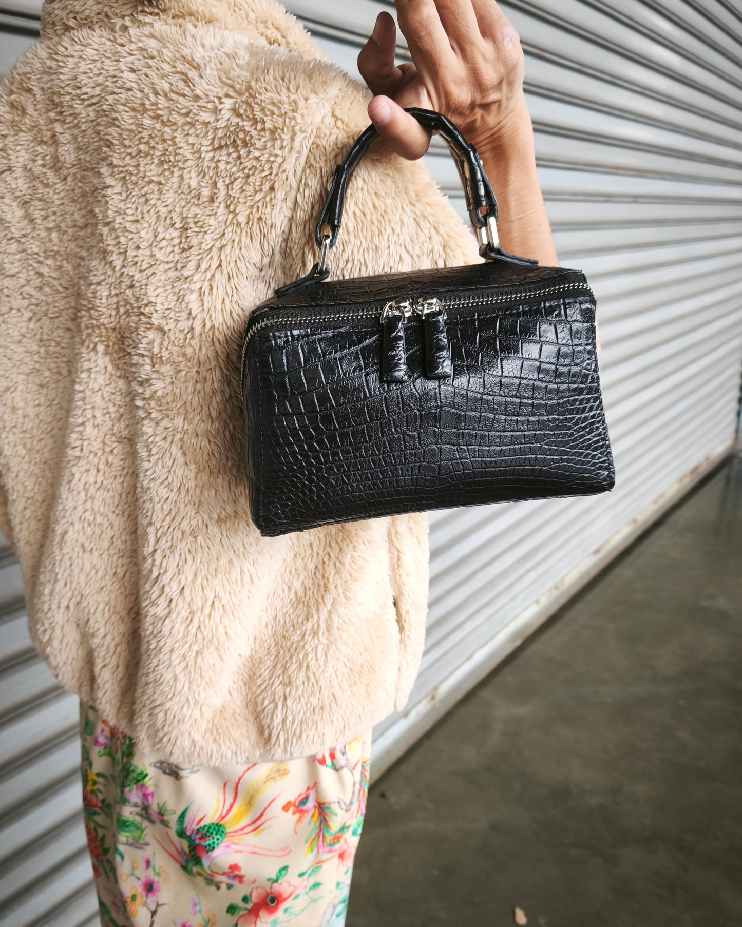 Person holding a black handbag with a textured surface, wearing a beige coat and floral pants.