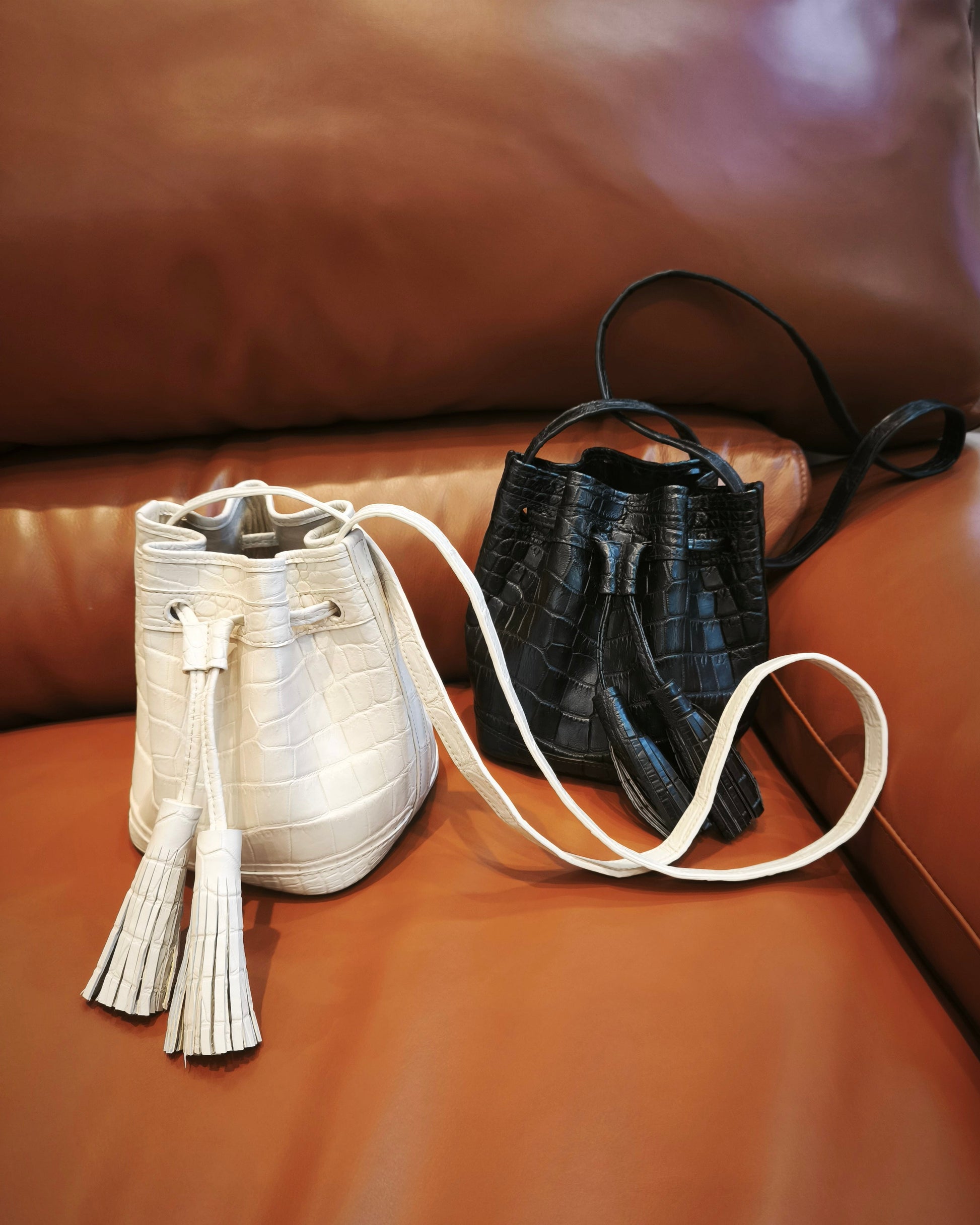 Two small bucket-style handbags placed on a brown leather sofa. The left bag is off-white/cream with a crocodile-embossed texture and a tassel drawstring.
